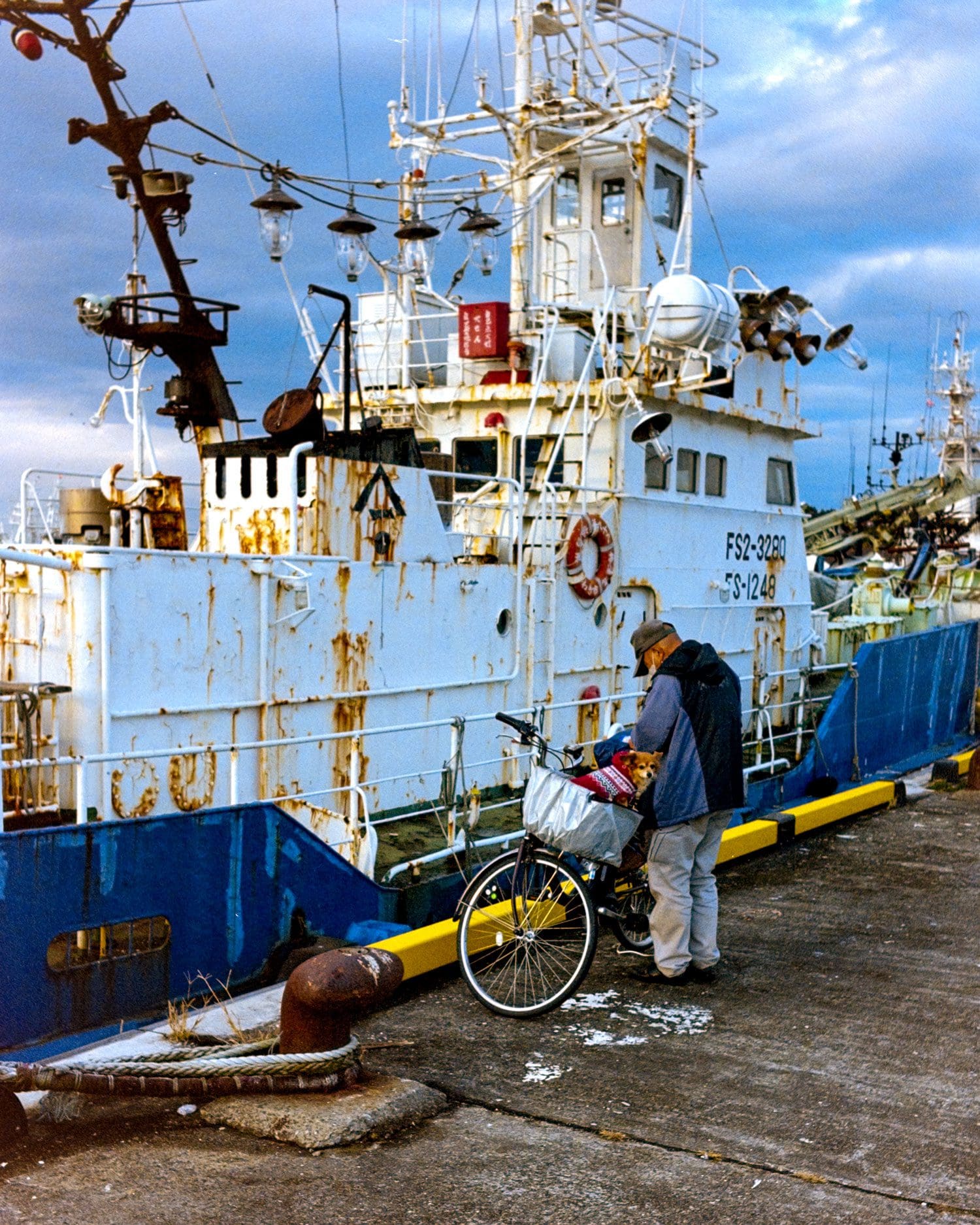 A man and his dog on a harbor dock with industry-grade fishing vessel backdrop under dramatic sky.