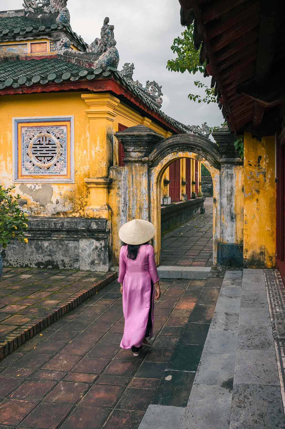Woman in traditional Vietnamese attire walking towards historic yellow archways in Historic Old Imperial Citadel of Hue .