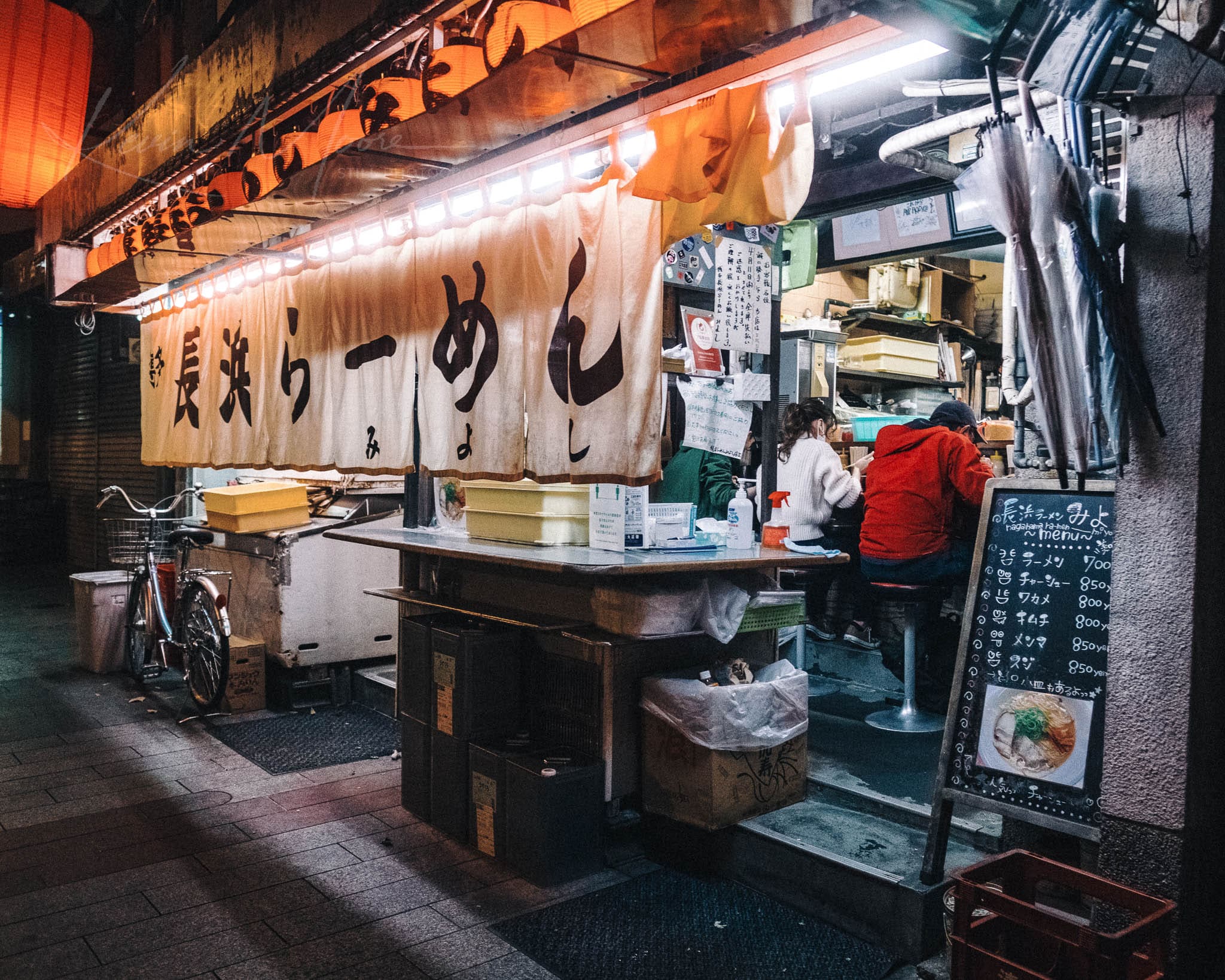 Vibrant night scene at an urban Kyoto ramen stall, packed with cooking tools and a bicycle.