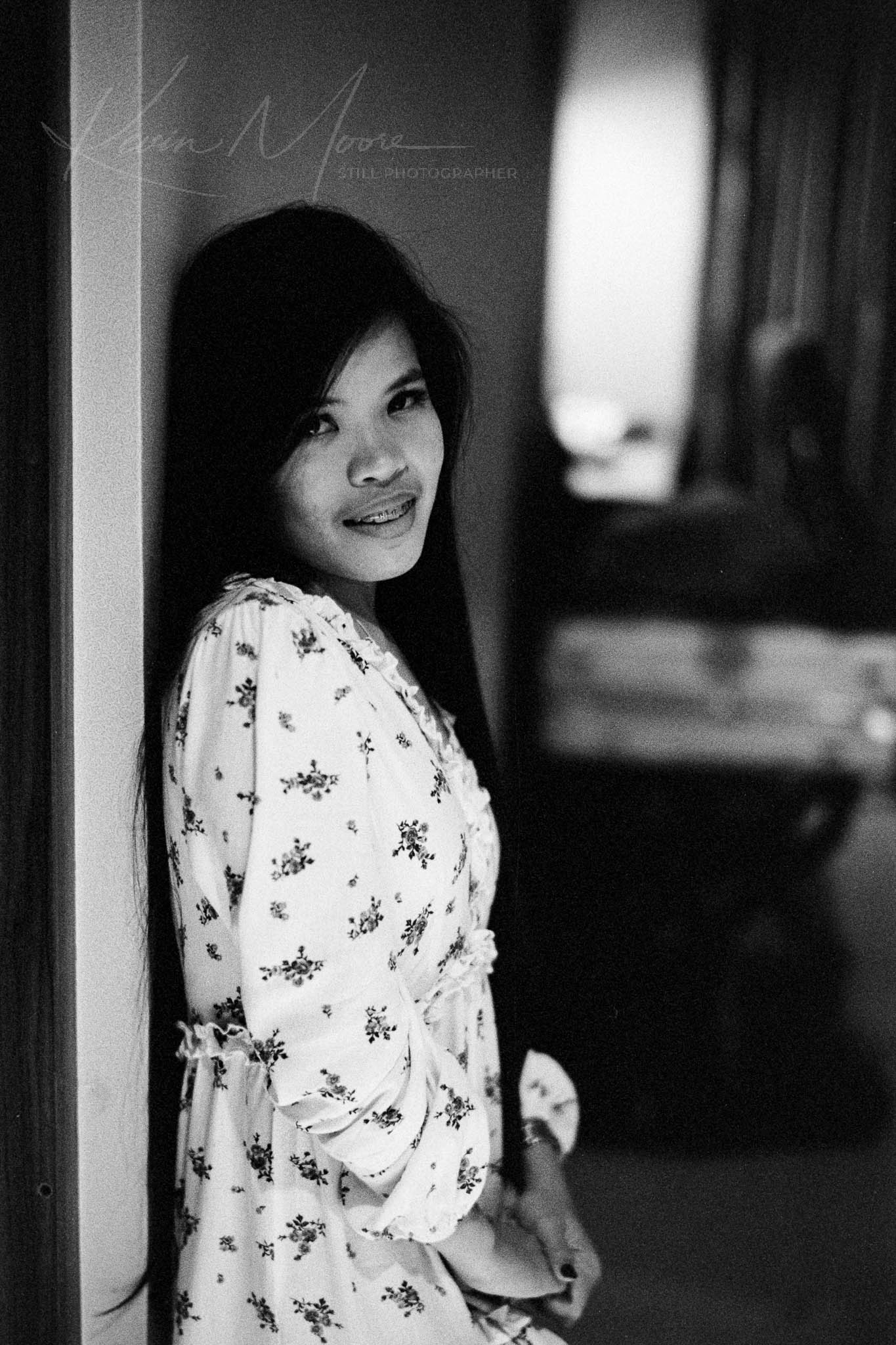 Monochromatic portrait of a smiling woman in floral dress leaning against a door frame.