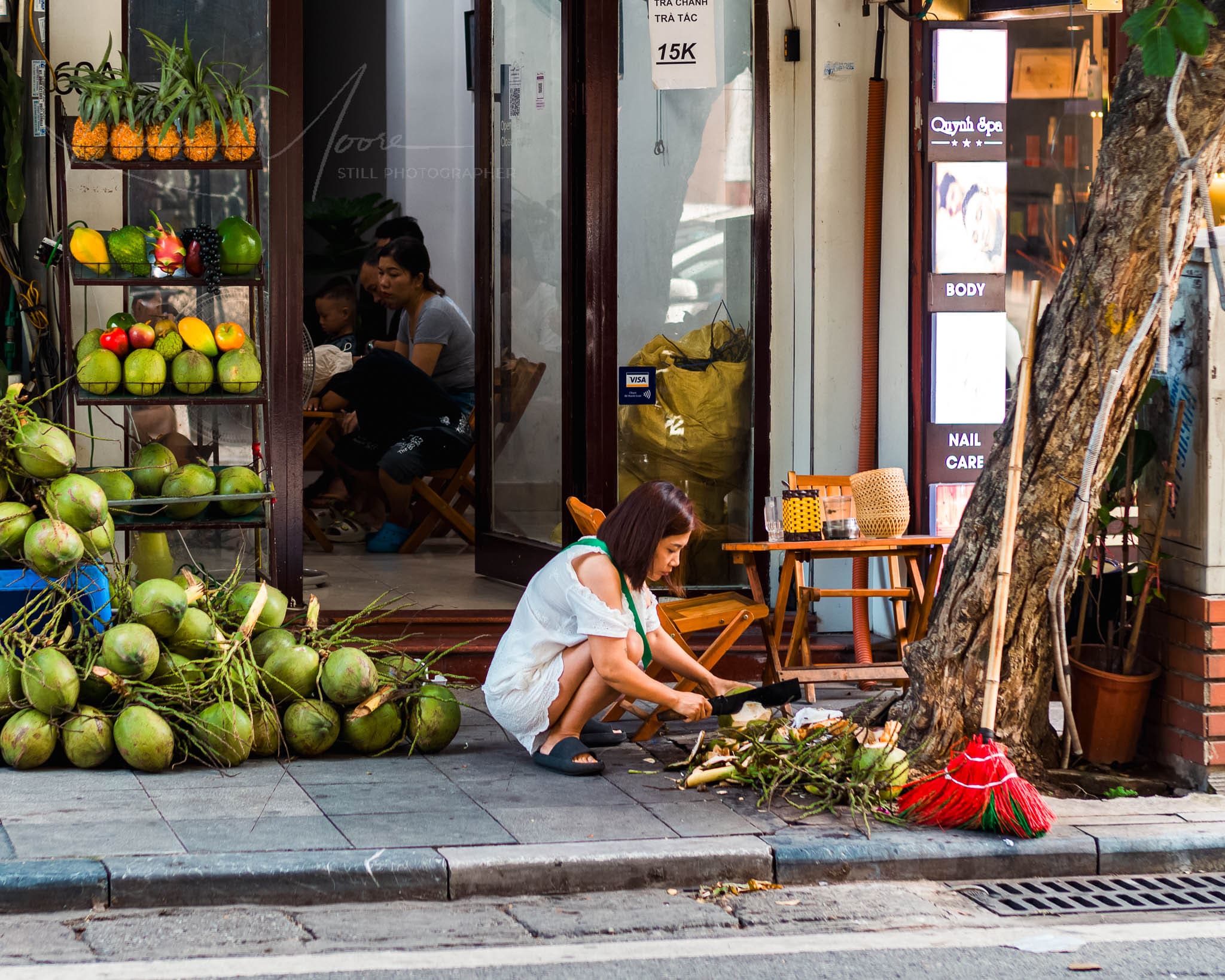 Tropical street scene with woman preparing coconuts, fresh fruit market, and urban tree.