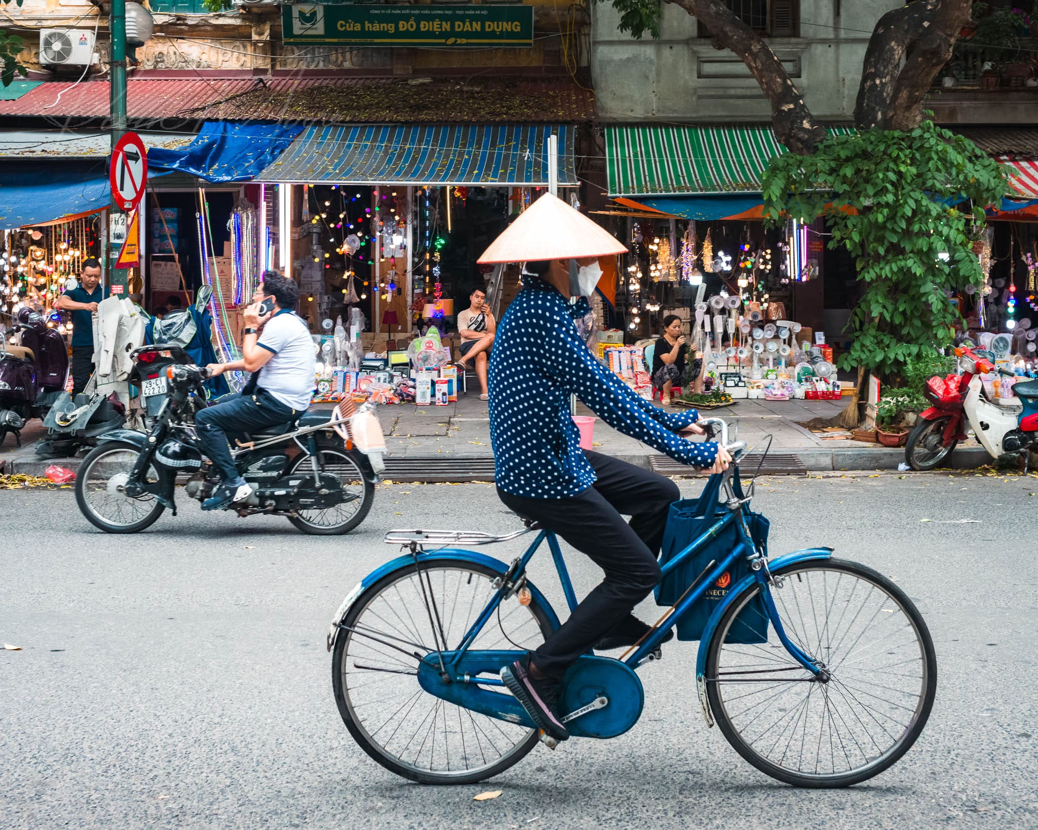 Cyclist riding on a bustling Hanoi city street with shops and motorcycles.