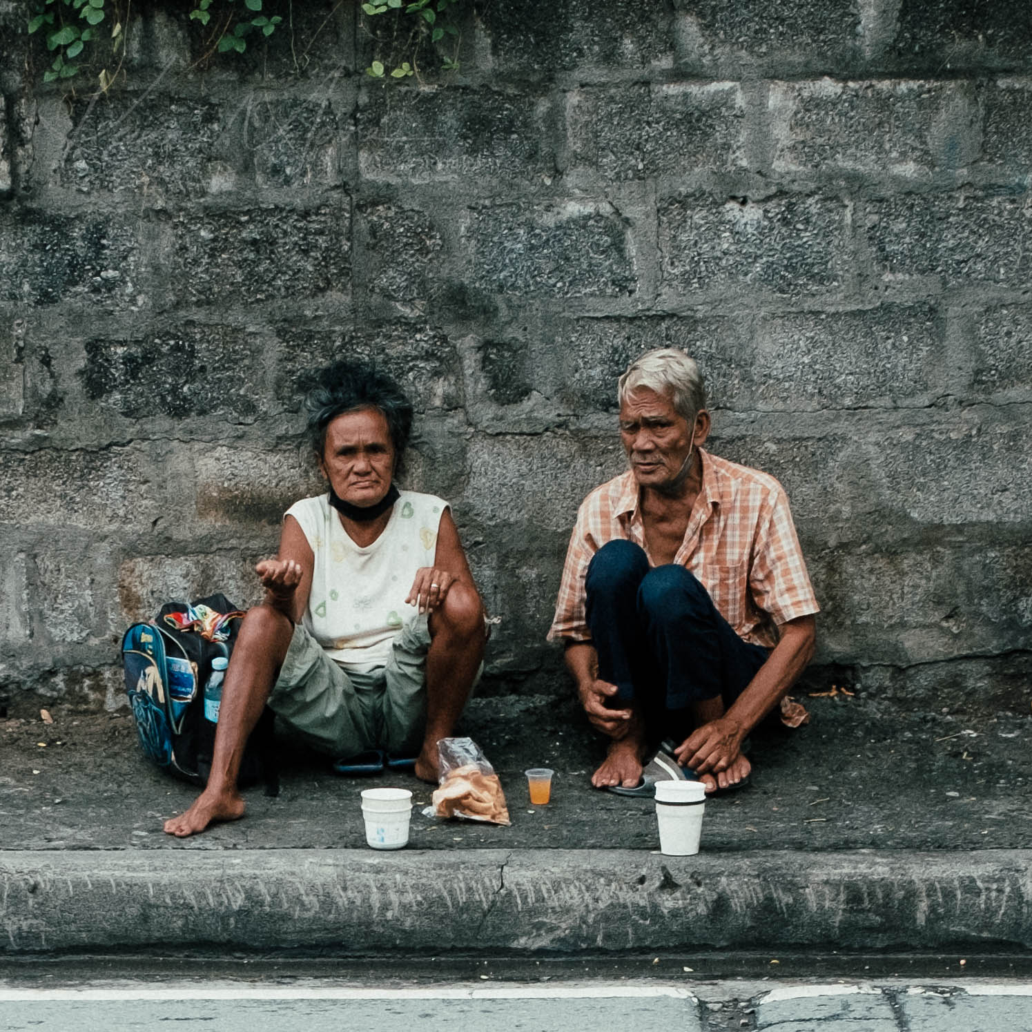 Elderly Filipino couple sitting roadside with coffee cups and personal belongings on a weathered ledge.