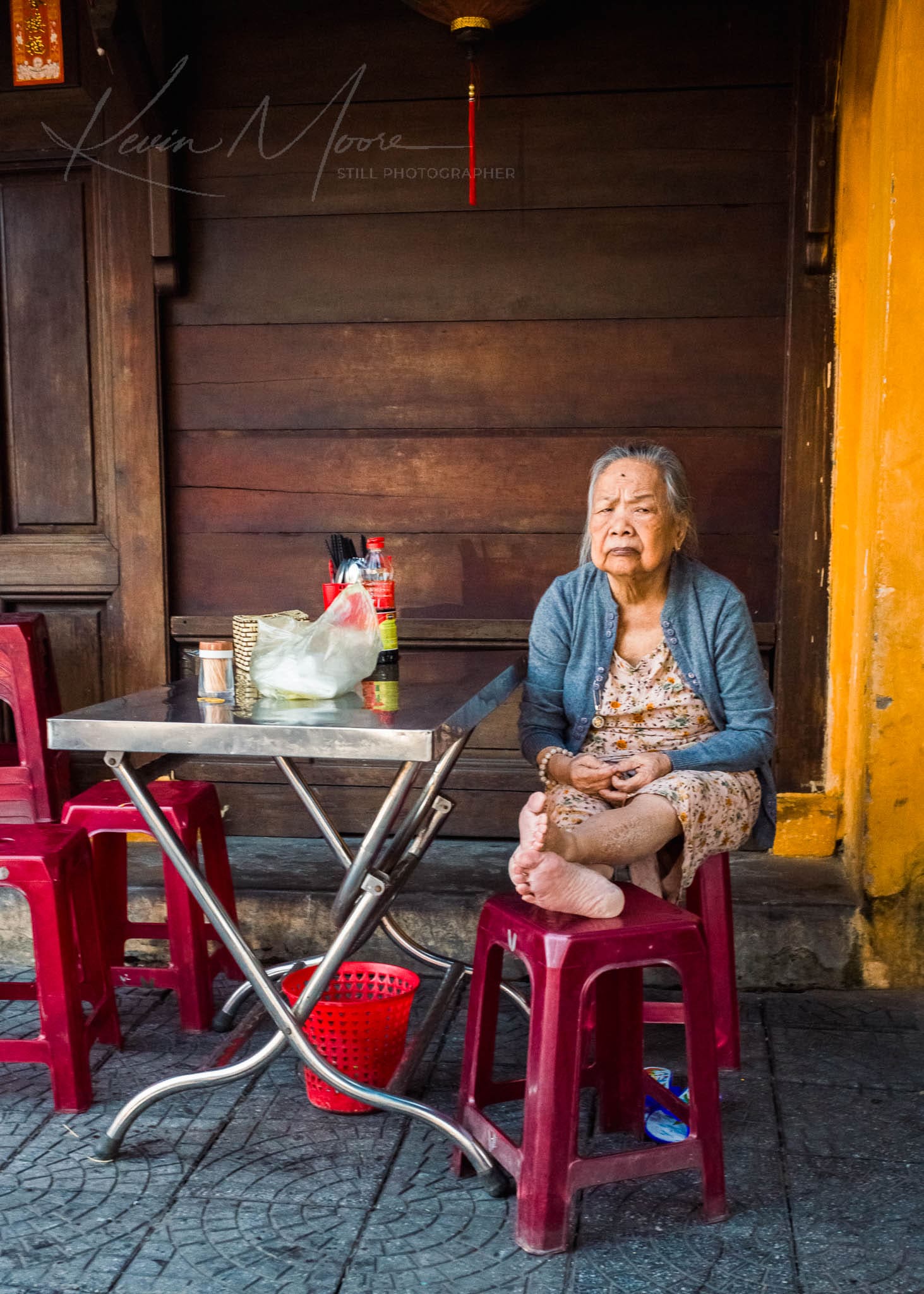 Elderly Vietnamese woman enjoying a peaceful moment in a rustic outdoor eatery during daytime.