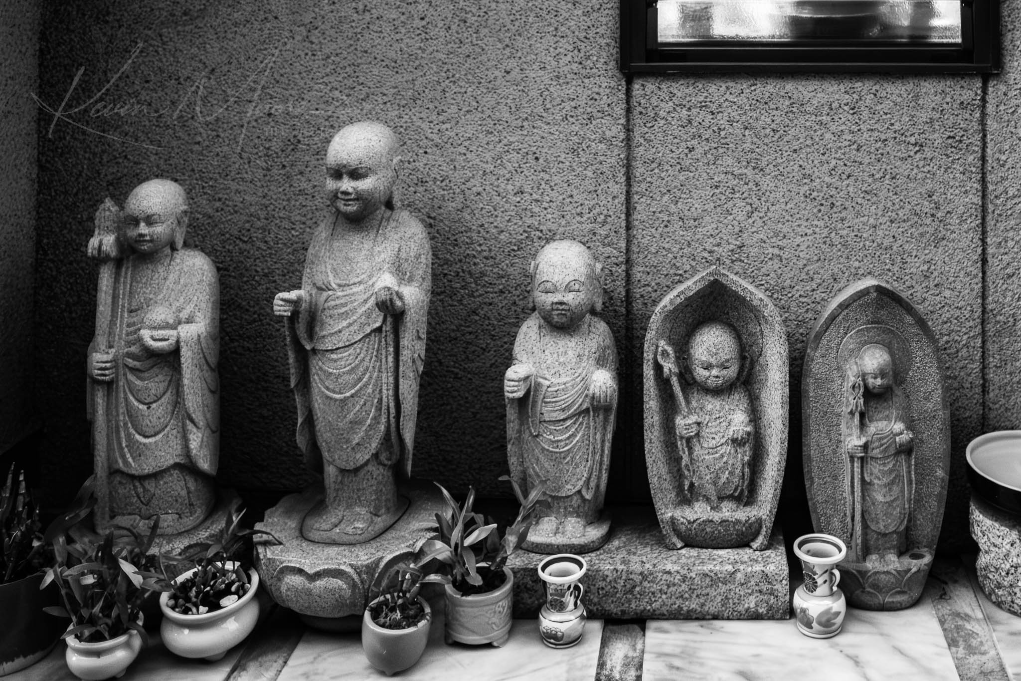 Monochrome image of five distinct Buddhist statues on a shelf with offerings.