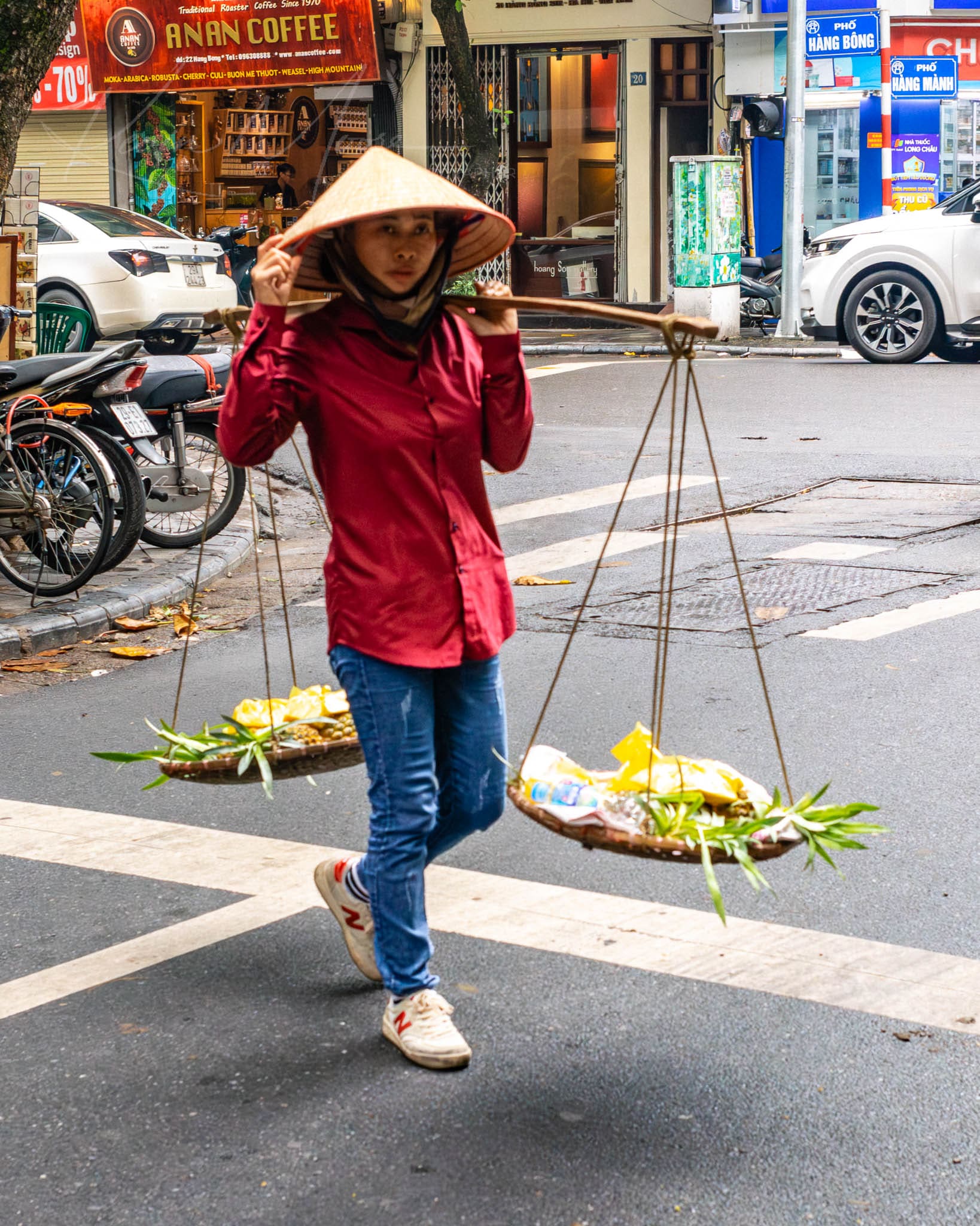 Woman carrying fresh produce with a yoke in an urban Hanoi Vietnam setting.