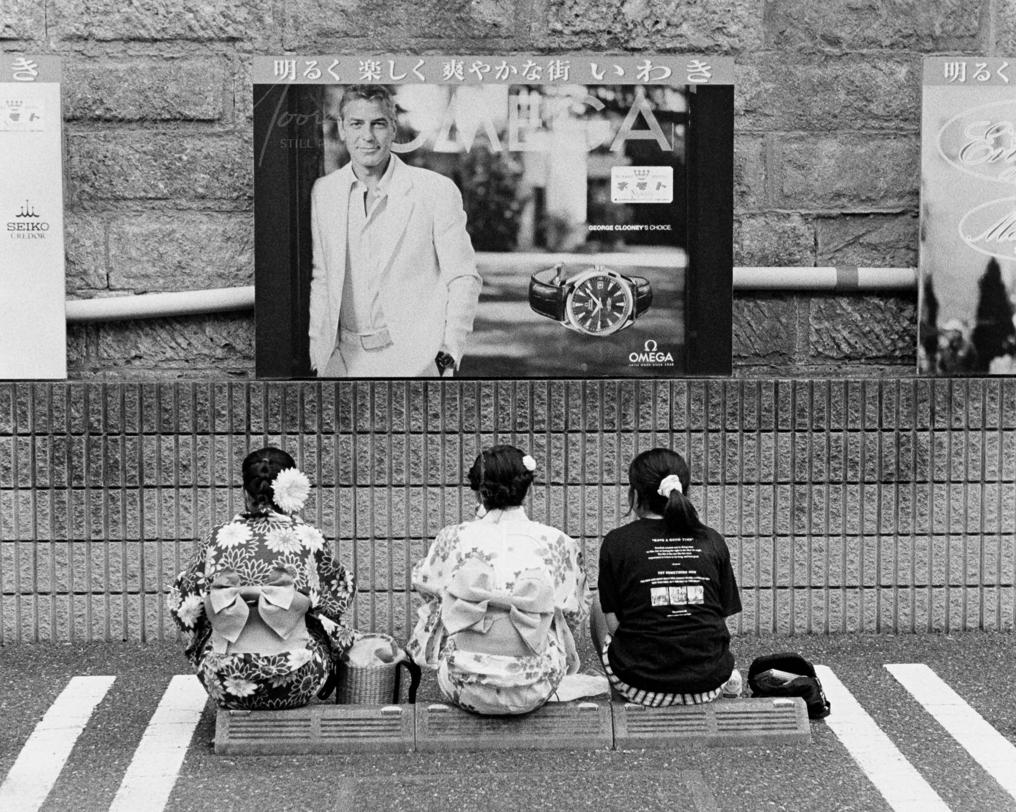 Three individuals in kimonos under a billboard of a man in Western attire, Japan.