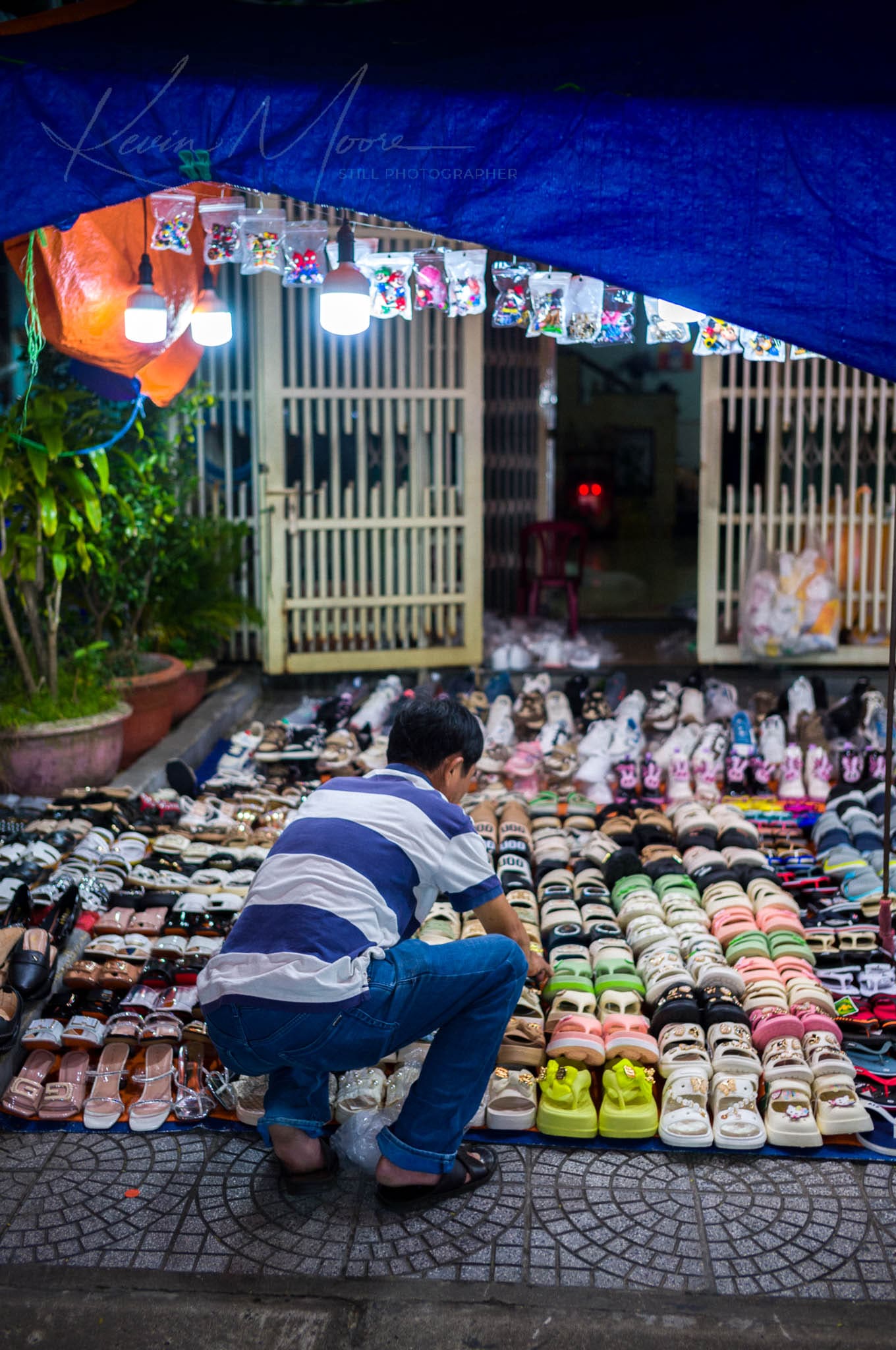 Nighttime shoe vendor organizing diverse footwear collection under a blue tarp at a street market.
