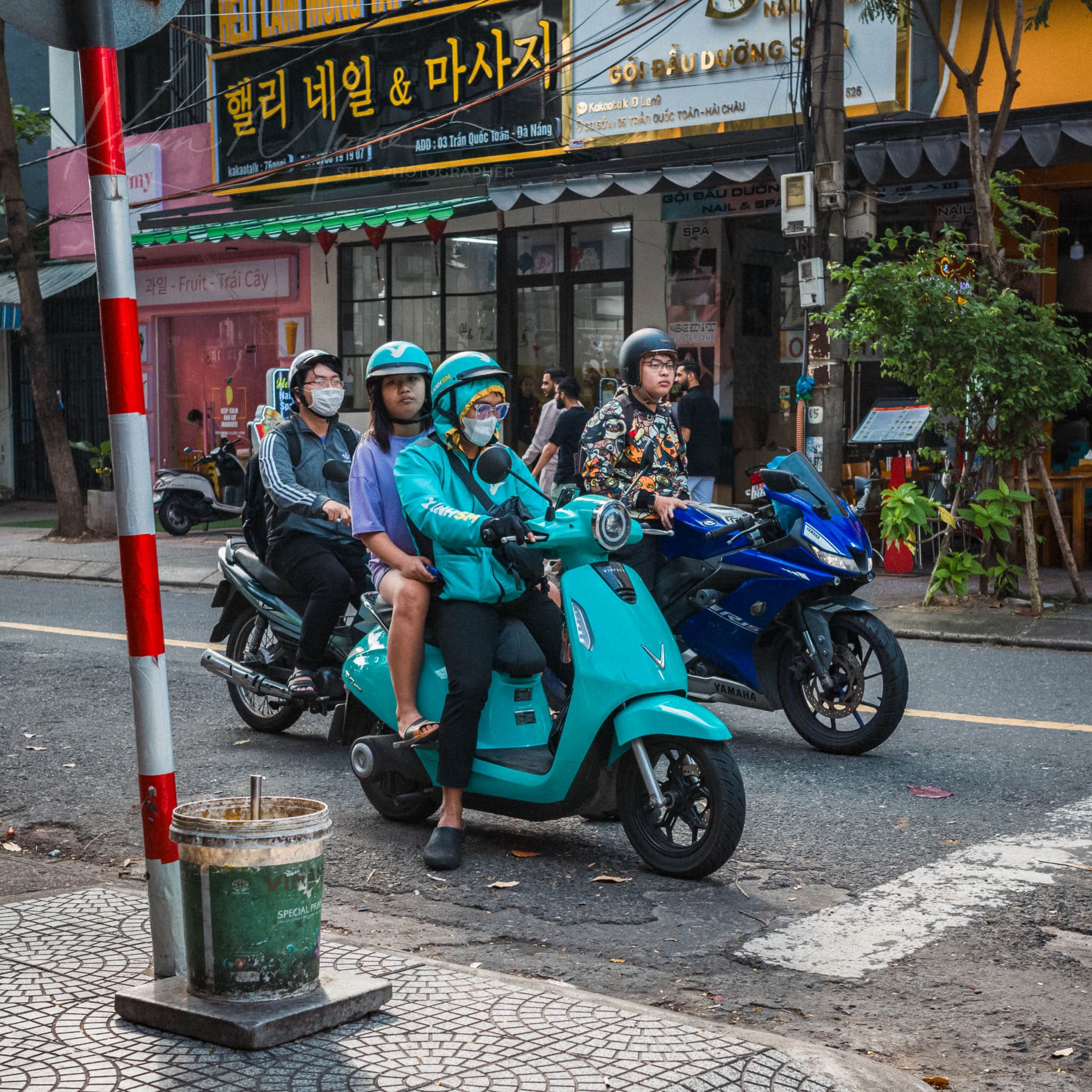 Four individuals waiting at a Da Nang city intersection on brightly colored scooters and motorcycles.