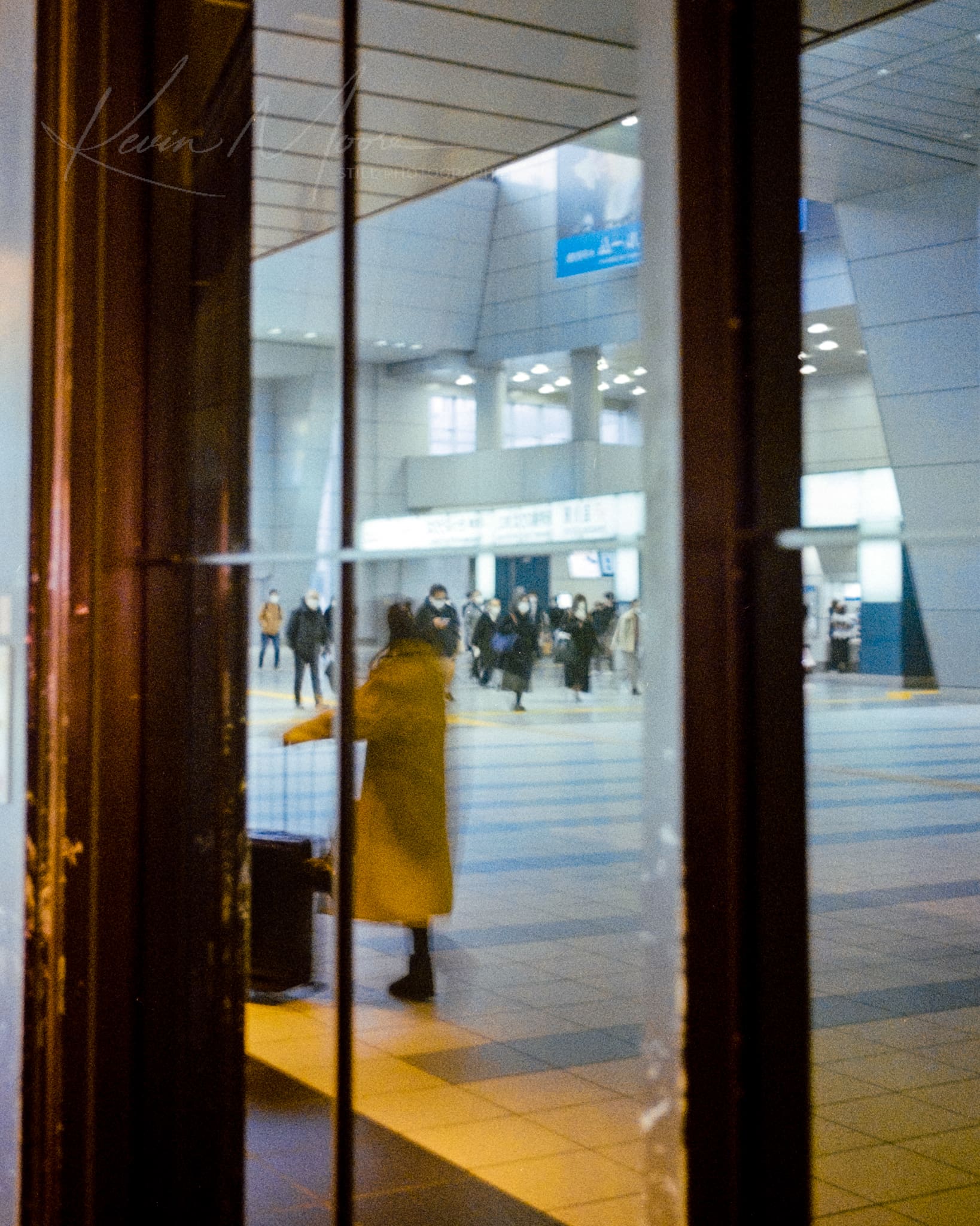 Woman in yellow coat at a bustling Tokyo transit station through glass reflection.