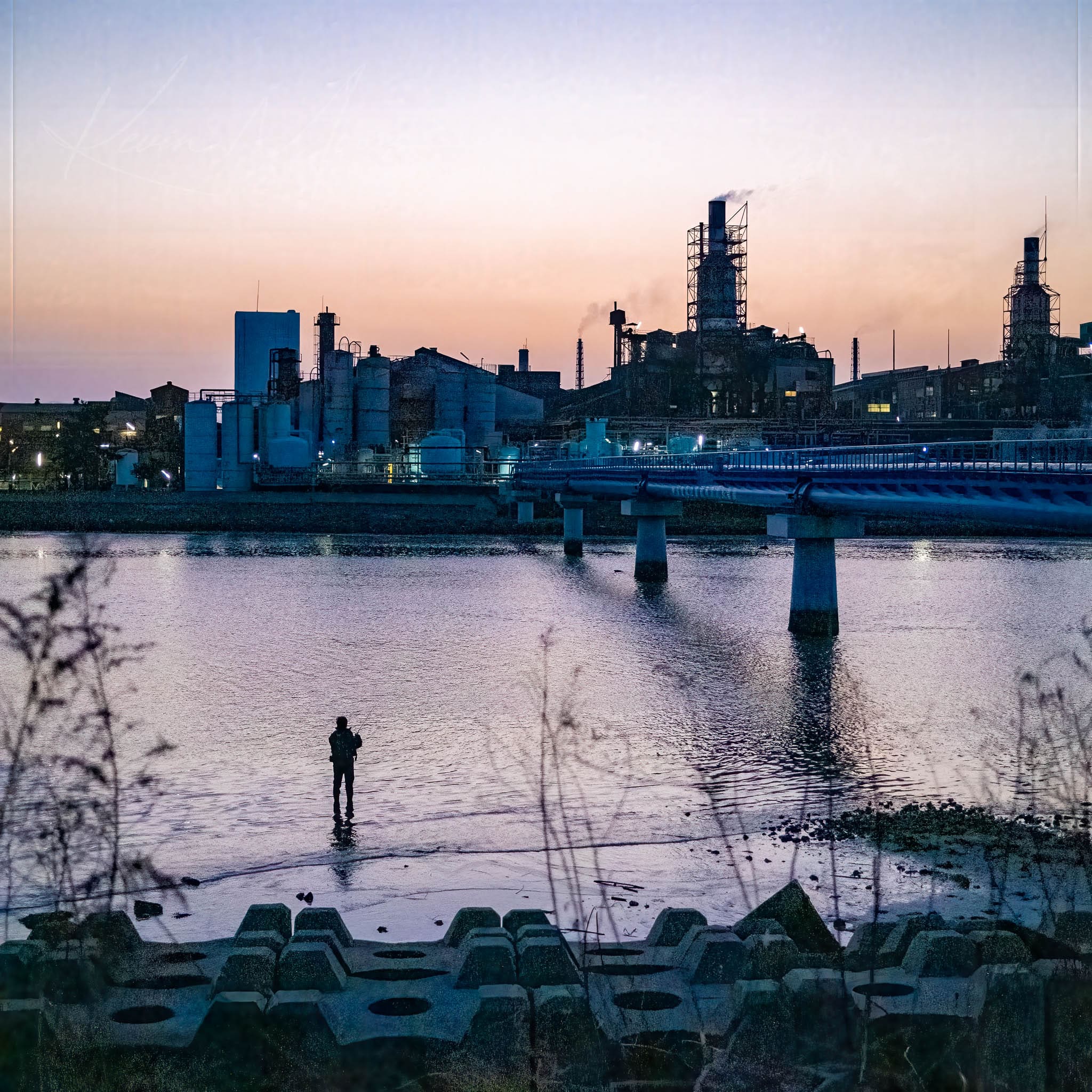 Solitary figure fishing by a serene inlet against a twilight industrial cityscape.