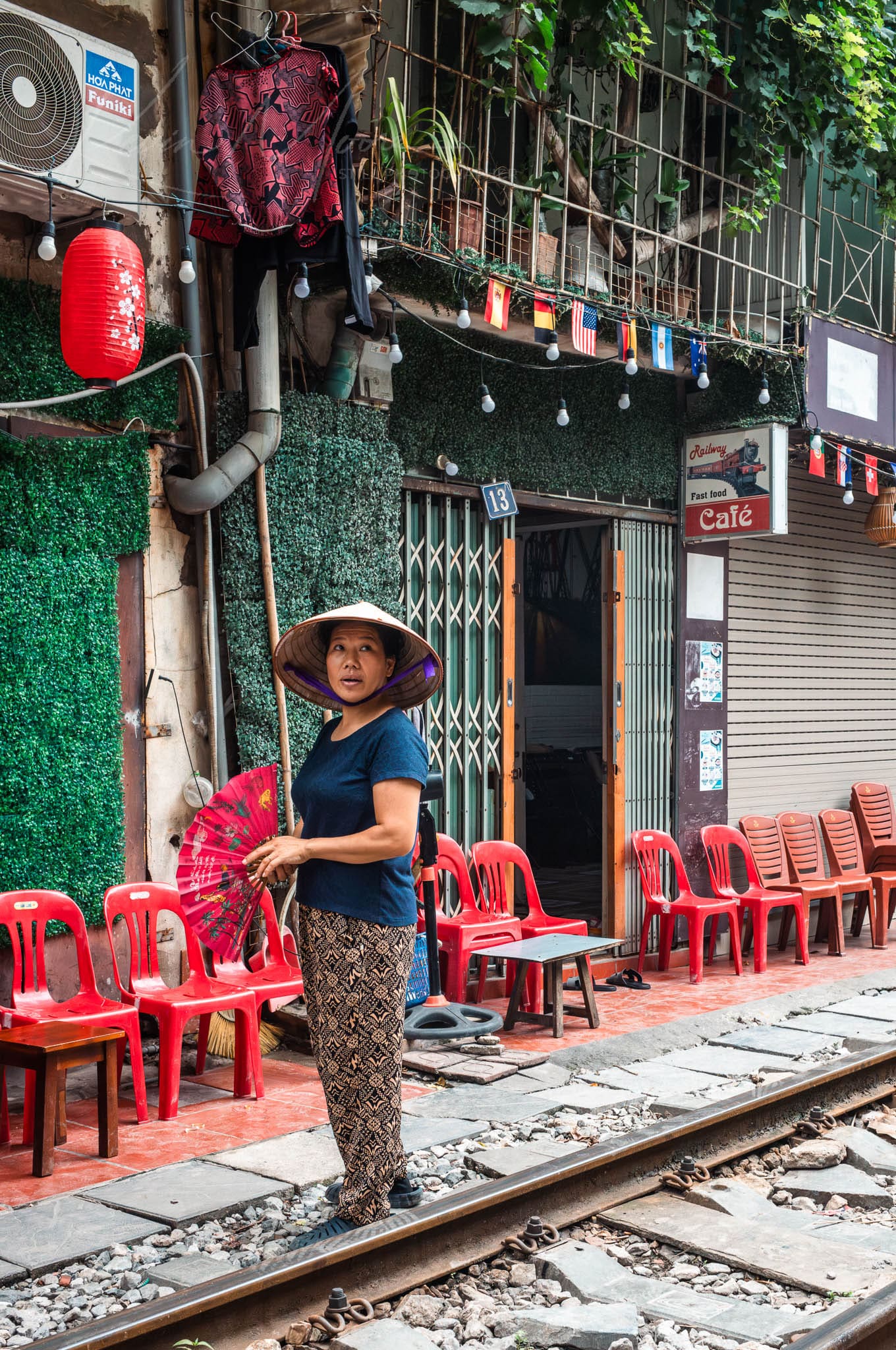 Vietnamese woman with traditional hat near urban Hanoi railway community with red chairs and lanterns.