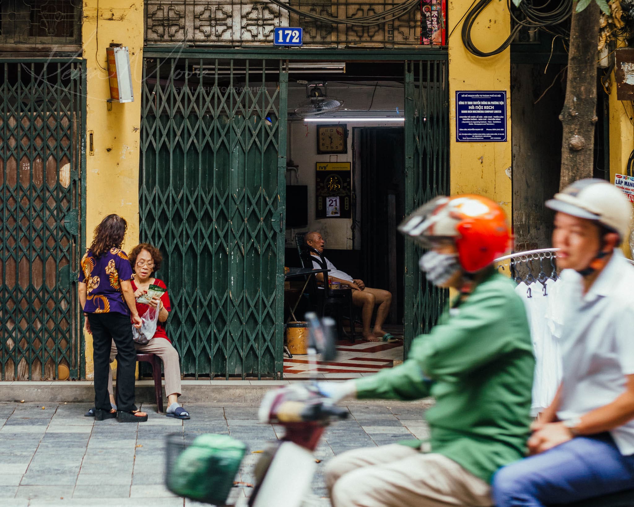 Motorbikes in motion and locals relaxing on a vibrant Hanoi Vietnam urban street scene.