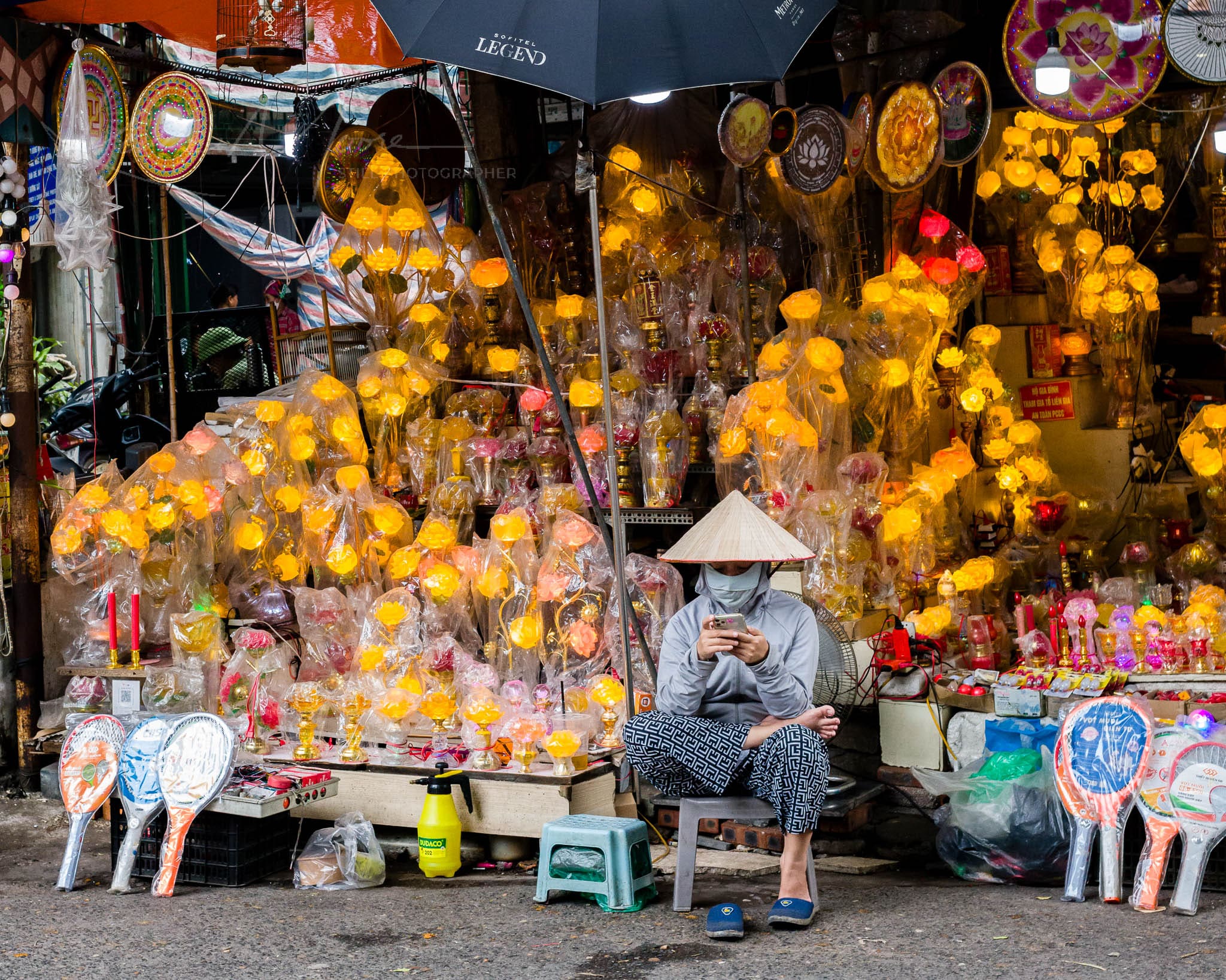Vendor in traditional Vietnamese attire using smartphone amid colorful handmade lanterns in a bustling market.