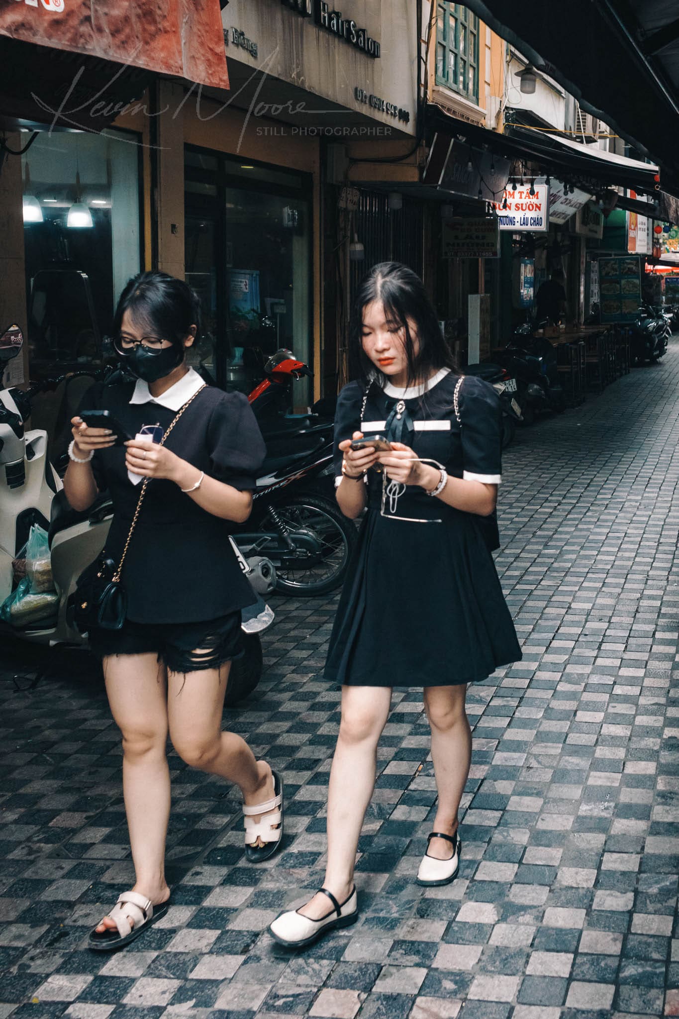 Two young Vietnamese women engrossed in smartphones on a tiled pedestrian street in Southeast Asia.