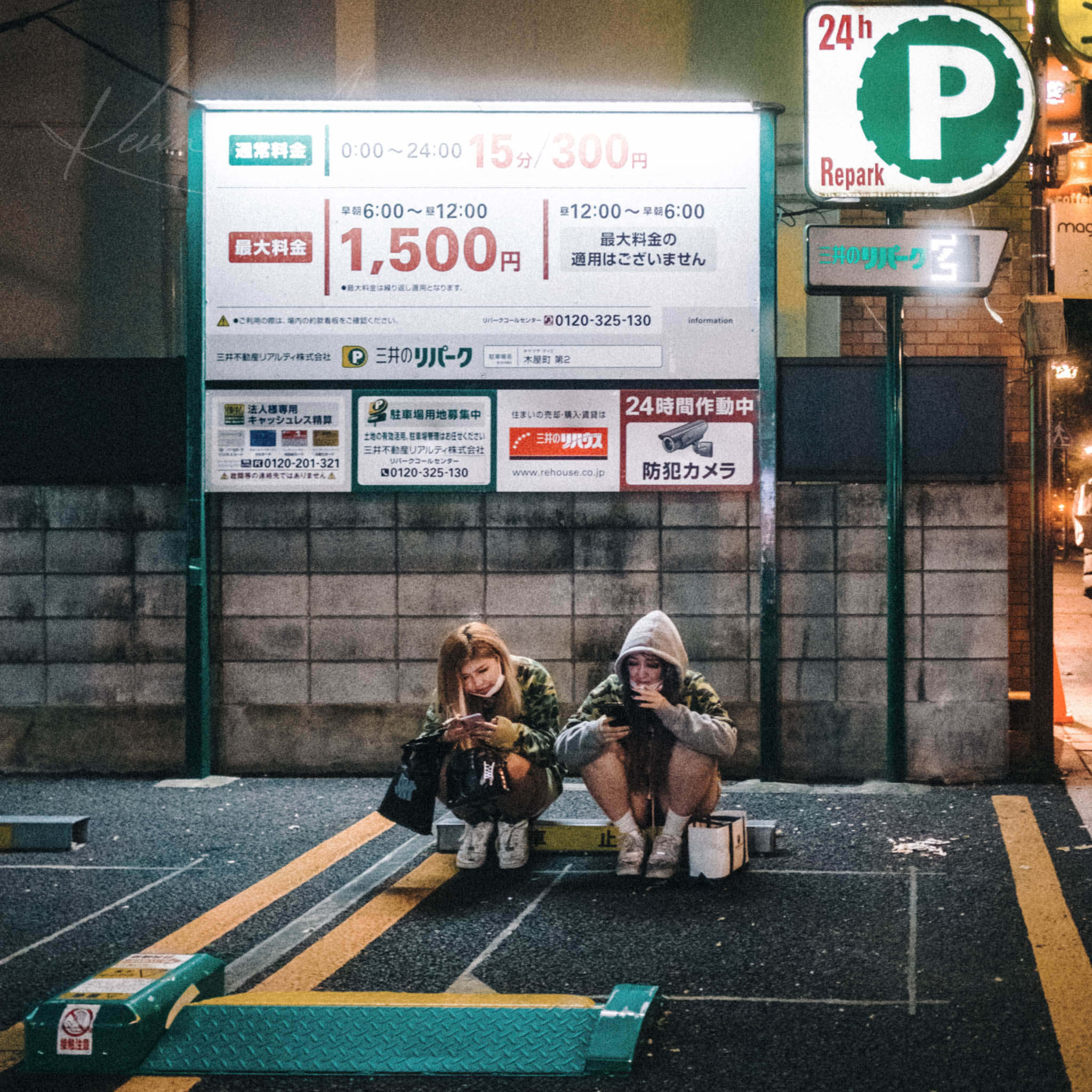 Two girls engrossed in smartphones at a lit-up 24-hour Repark parking lot in Kyoto at night.