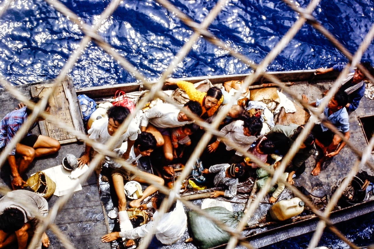 Overcrowded boat filled with Vietnamese refugees amid deep blue sea, viewed through mesh barrier.