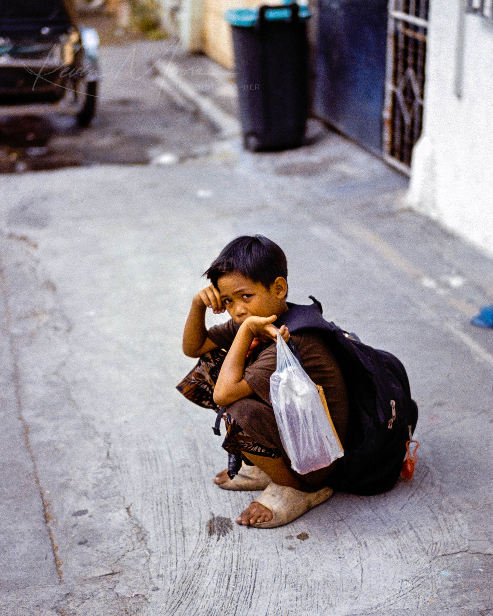 Young boy reflecting on urban street, holding plastic bag, under warm afternoon light.