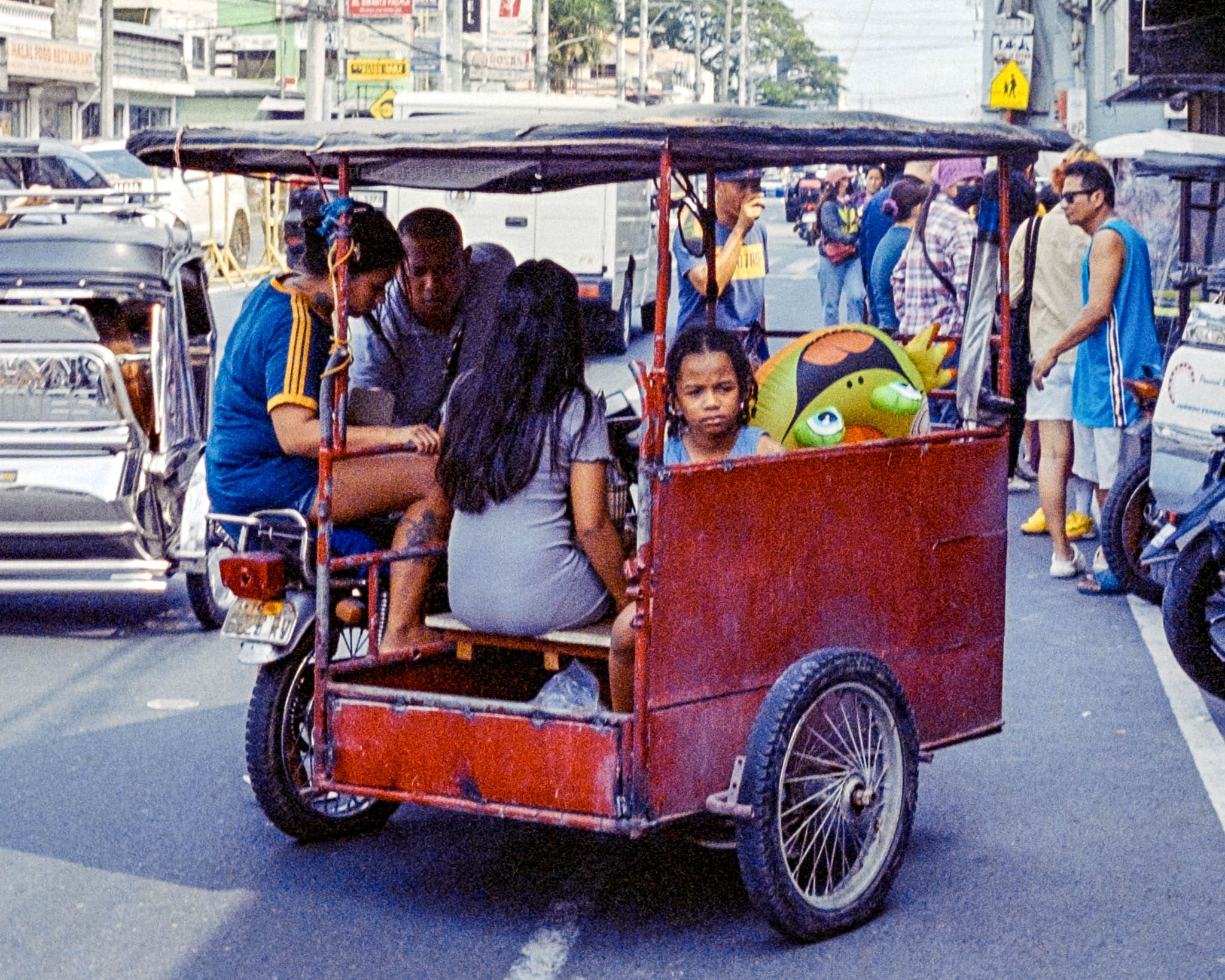 Family and stuffed bird on a trike amidst bustling Southeast Asian city street activities.
