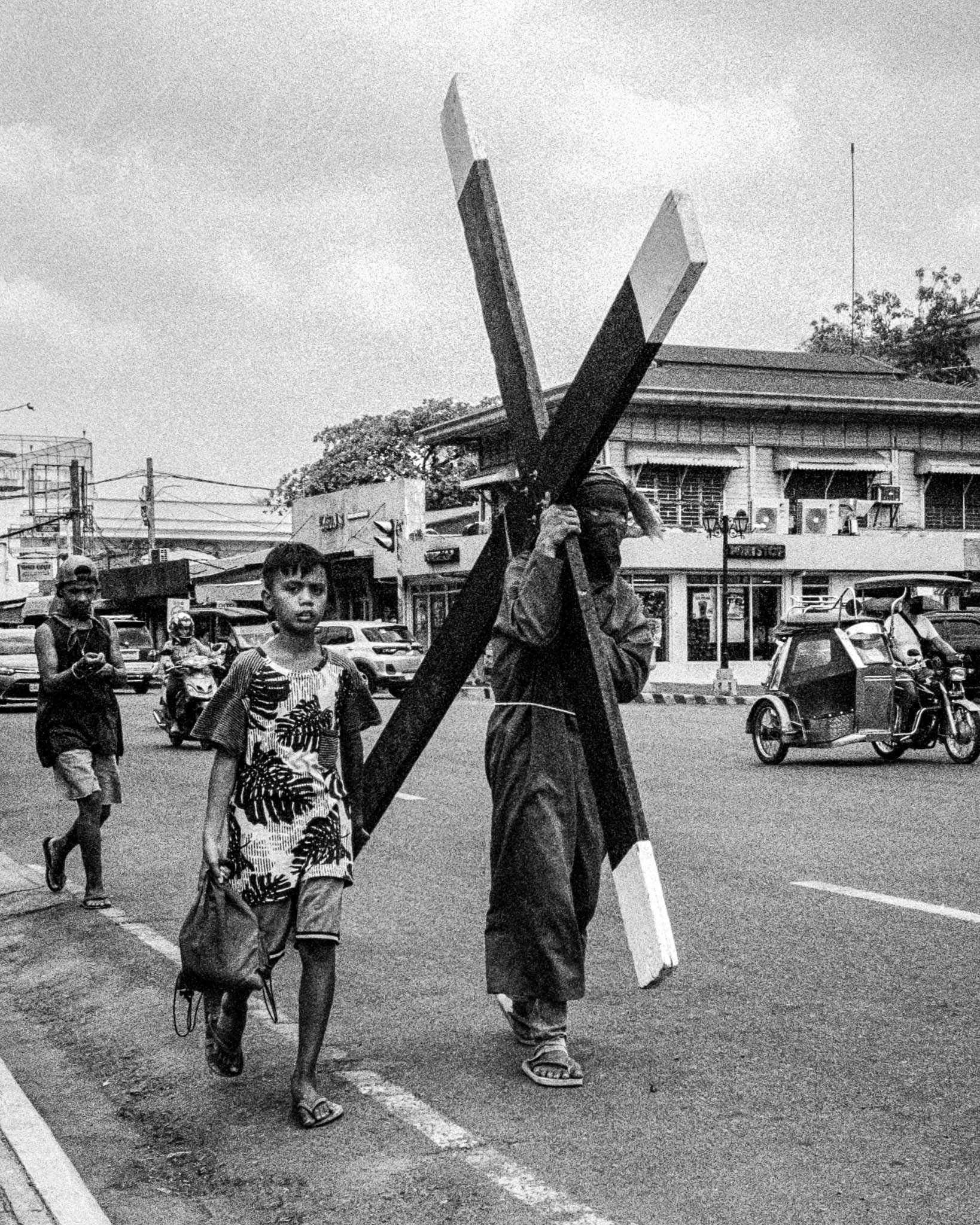 Man carrying large wooden cross in religious ceremony on city street, accompanied by child.