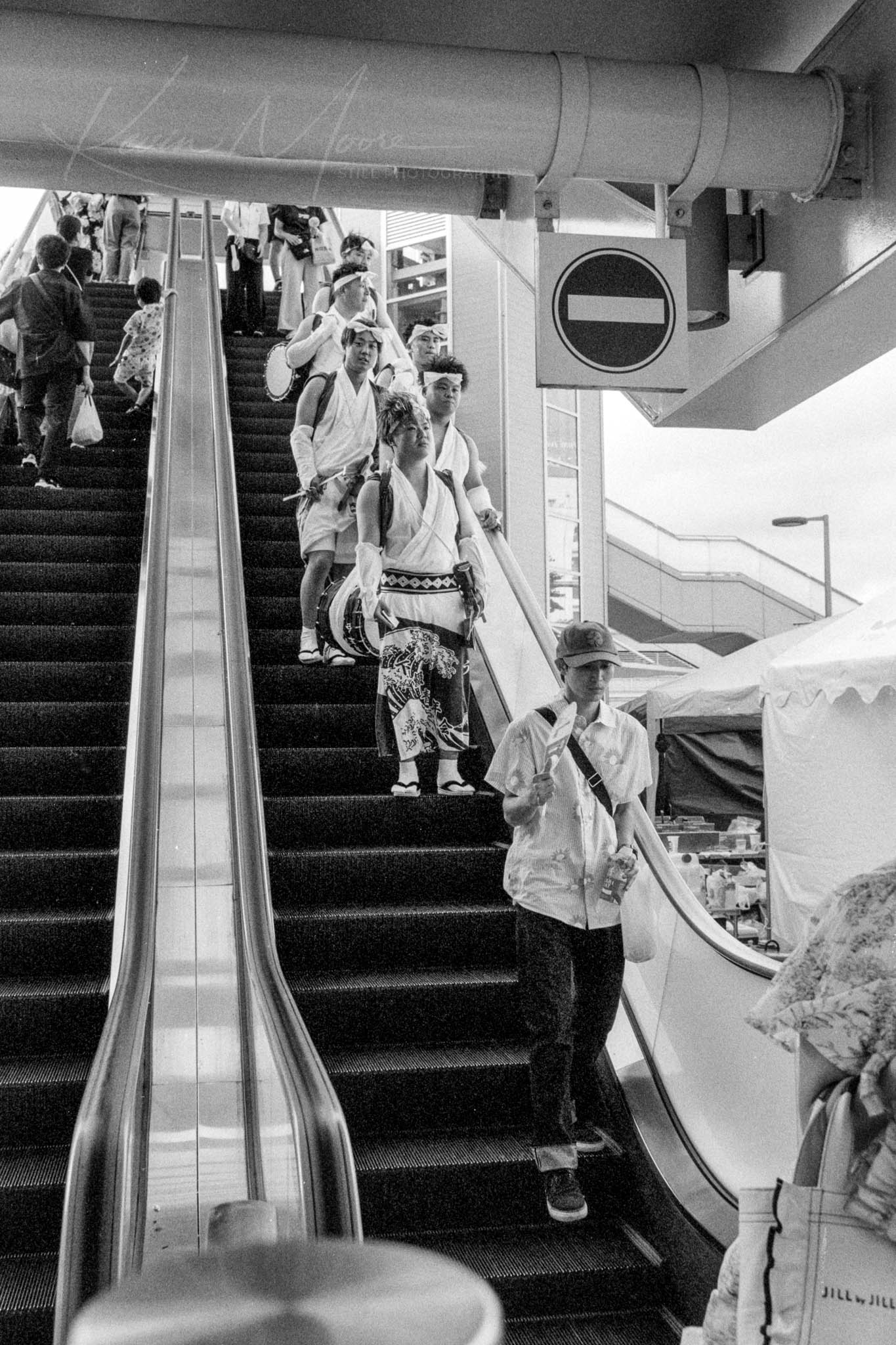 Candid black and white photo of Japanese Jangara performers on an descending escalator.