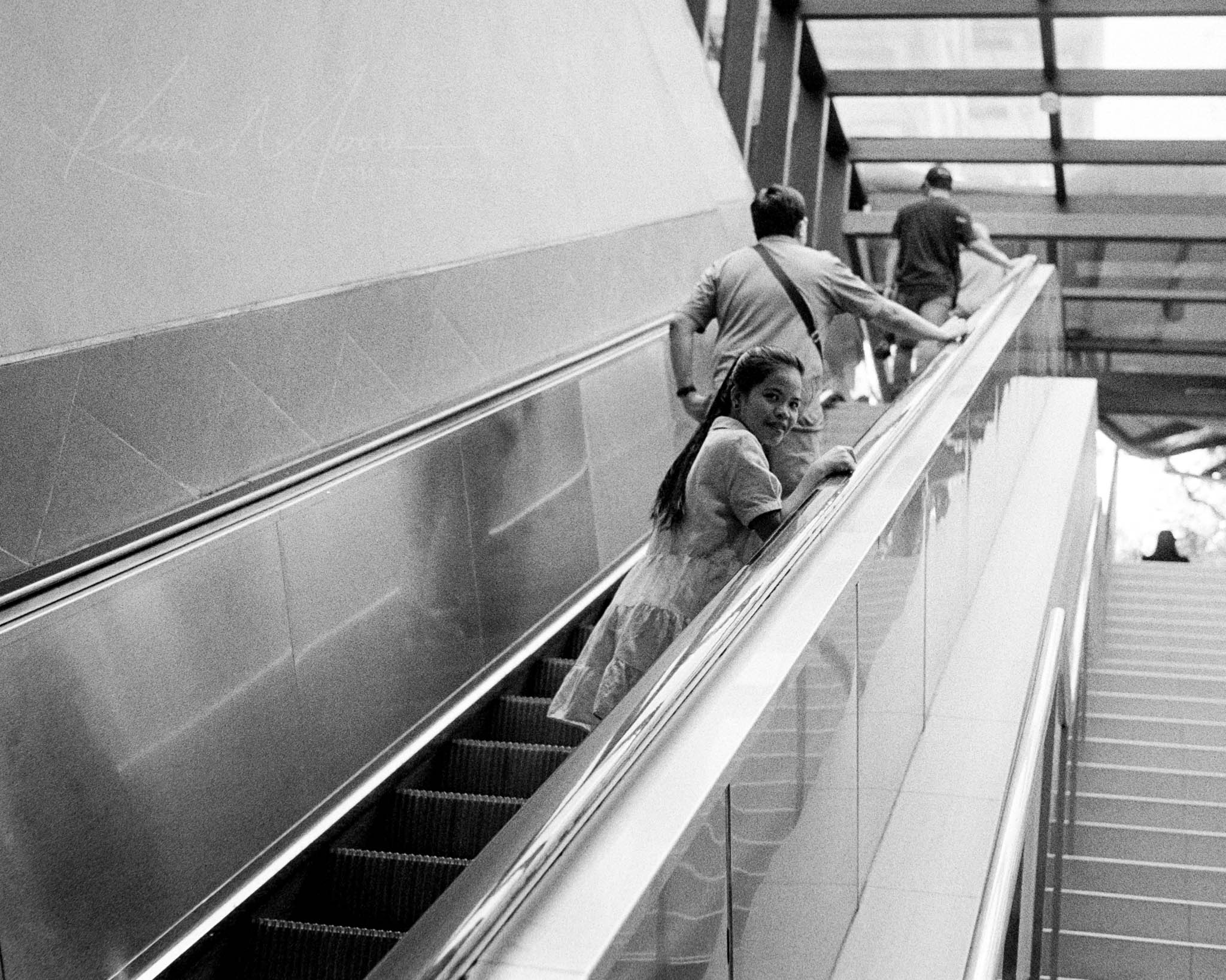 Monochrome image of people on escalator, with young woman engaging curiously with the camera.