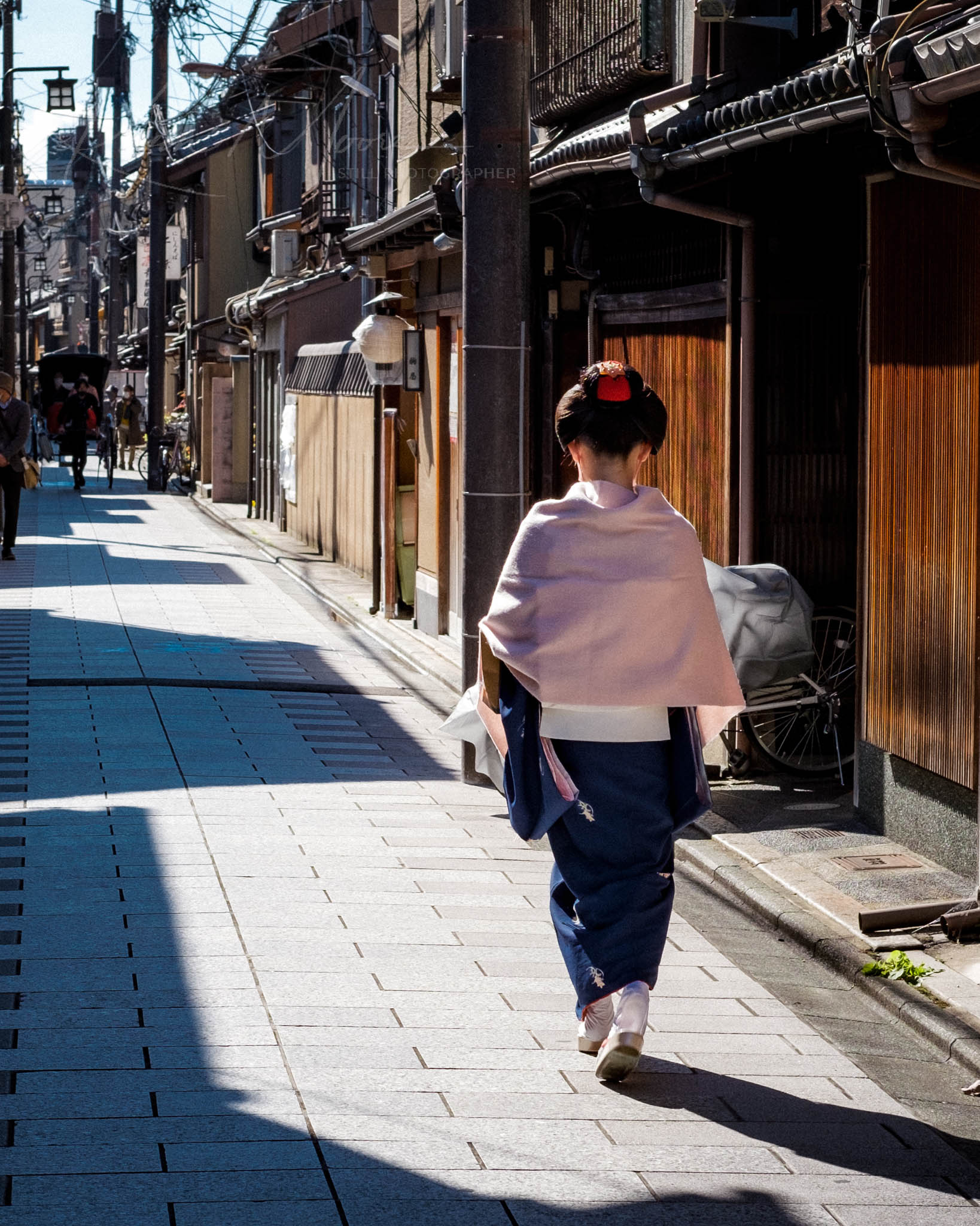 Geisha in kimono strolling on sunlit, traditional cobblestone street in Kyoto, Japan.
