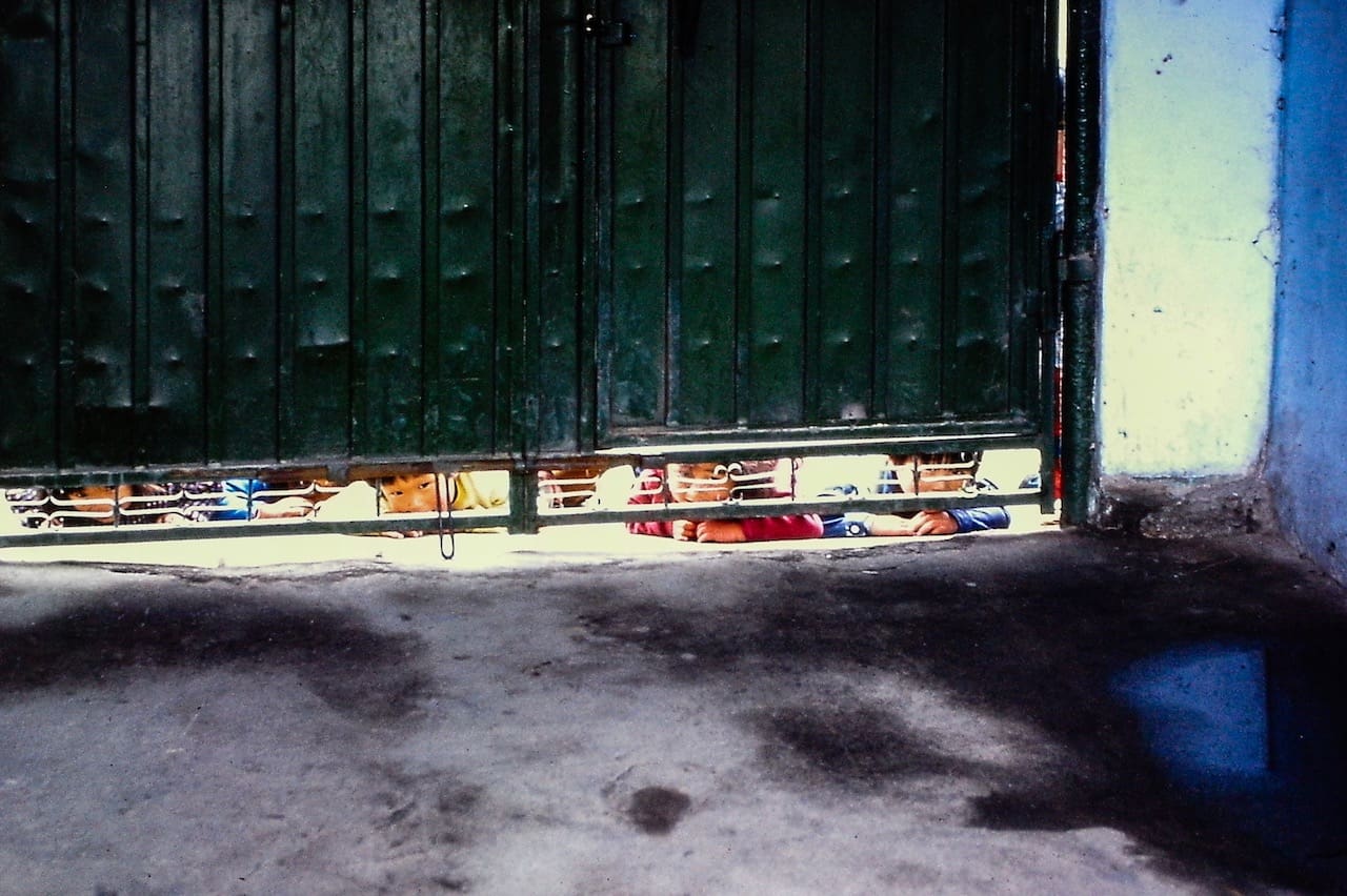 Curious Korean children peeking from beneath an aged green metal gate in a vibrant urban setting.
