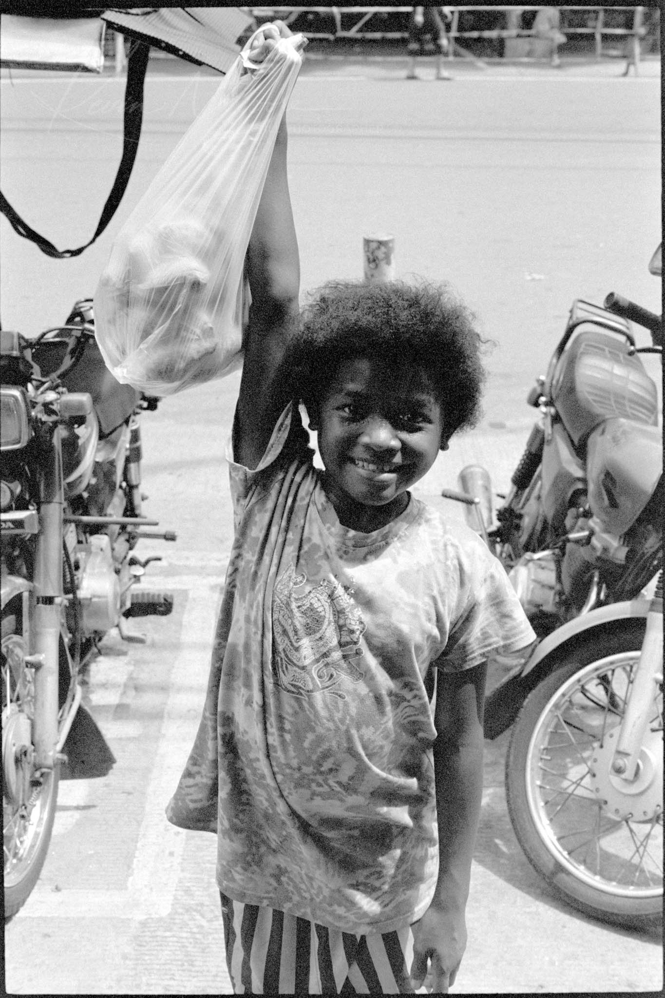 Smiling Child Holding Bag in Sunny Urban Street, Black and White Photo