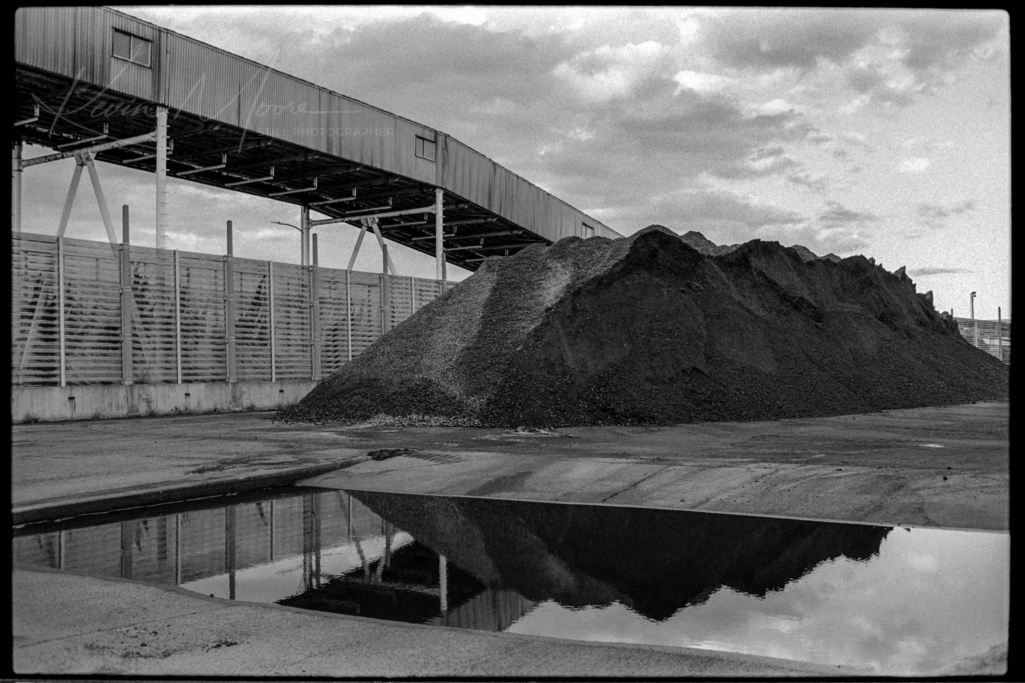 Black and white image of an idle industrial yard with gravel pile and mirrored reflections.