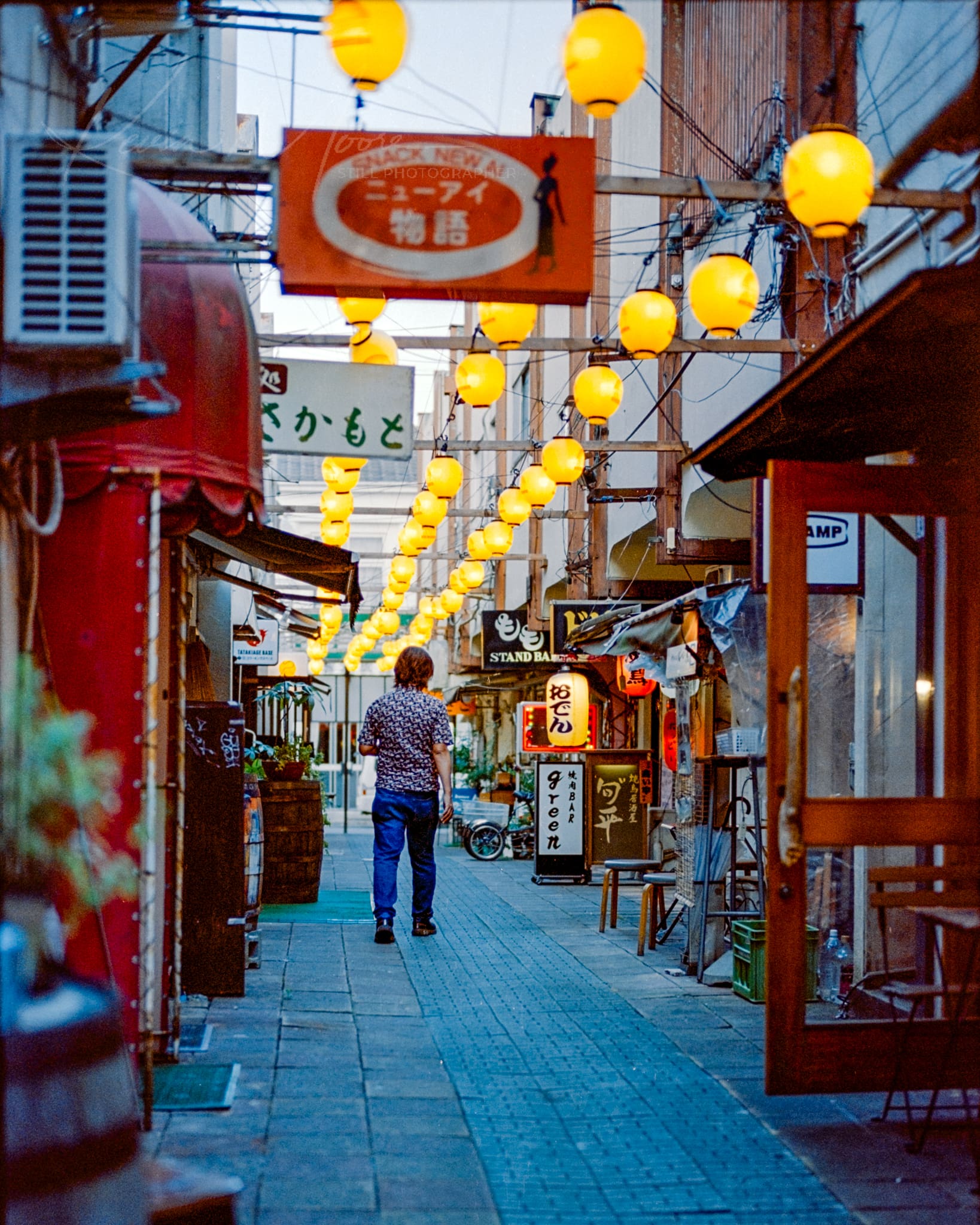 Japanese alleyway at dusk featuring traditional lanterns, eclectic storefronts, and a lone pedestrian.