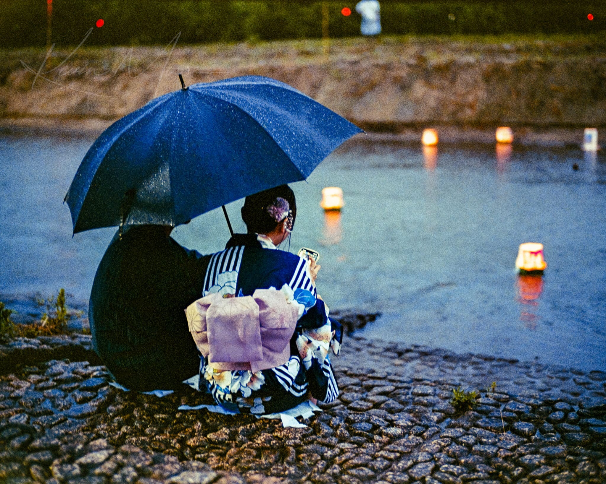 A couple in traditional Japanese attire sitting by a river with floating lanterns at twilight.
