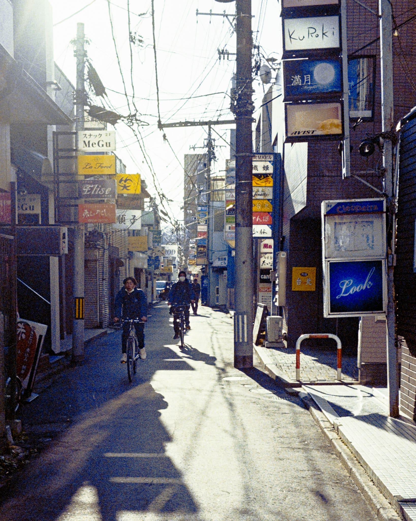 Sunlit Japanese street with cyclist and pedestrians amidst shadows in a serene urban scene.