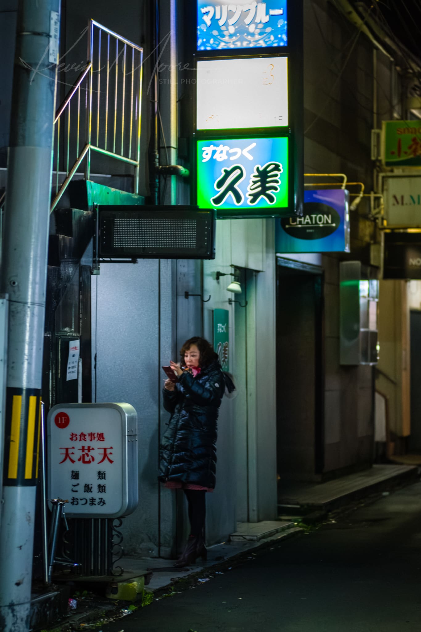 Woman using smartphone on rainy night in Japanese cityscape lit by neon signs.