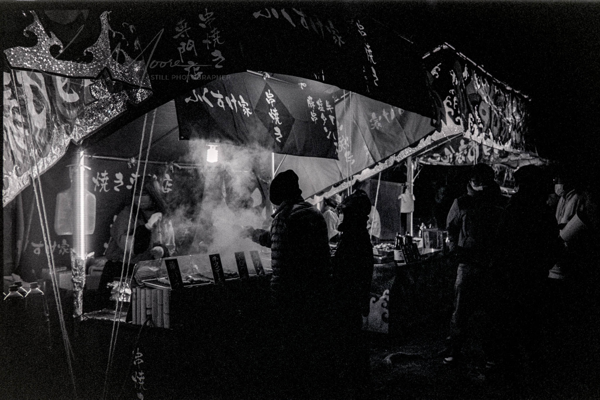 Bustling night market scene with illuminated food stall and customer silhouettes in black and white.