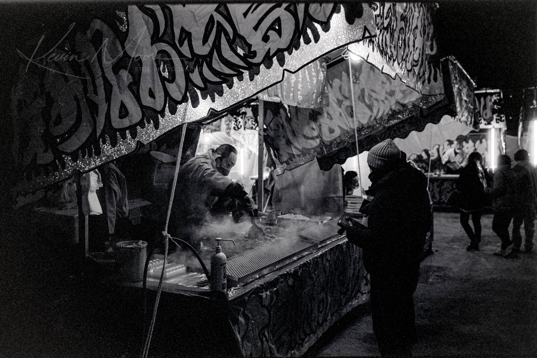 Bustling night market scene in Japan with illuminated, intricately adorned stall and smokey ambiance in black and white.