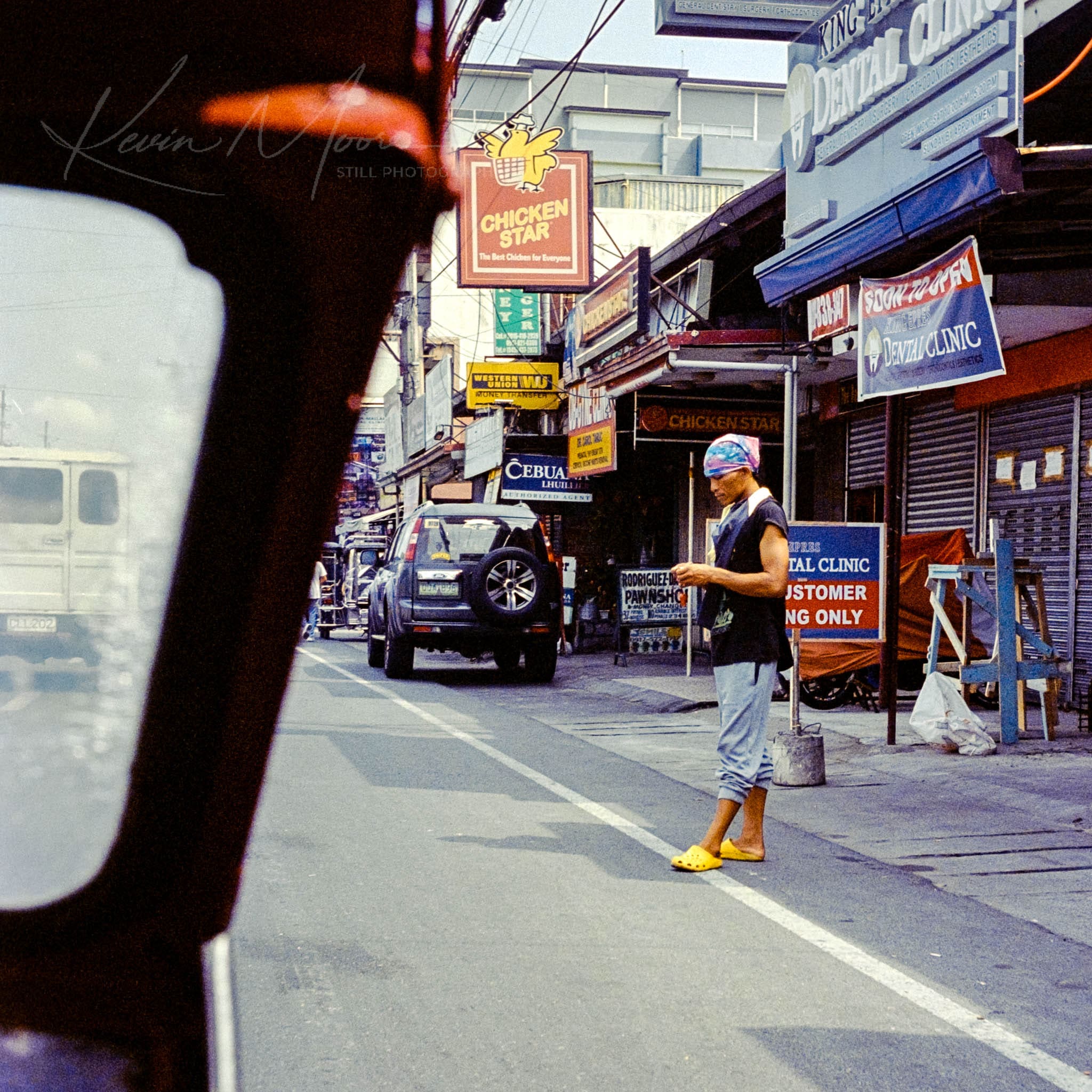Person in mask using phone on a quiet urban street with English storefronts and parked vehicles.