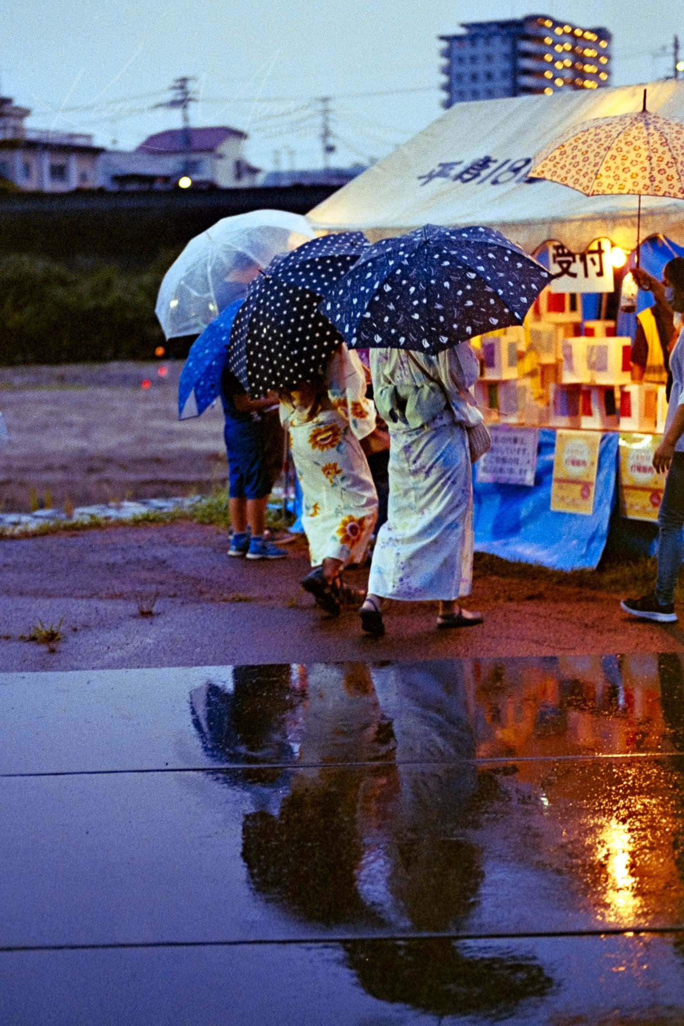 Group under colorful umbrellas on rainy evening, reflecting on wet ground near a lit food cart.