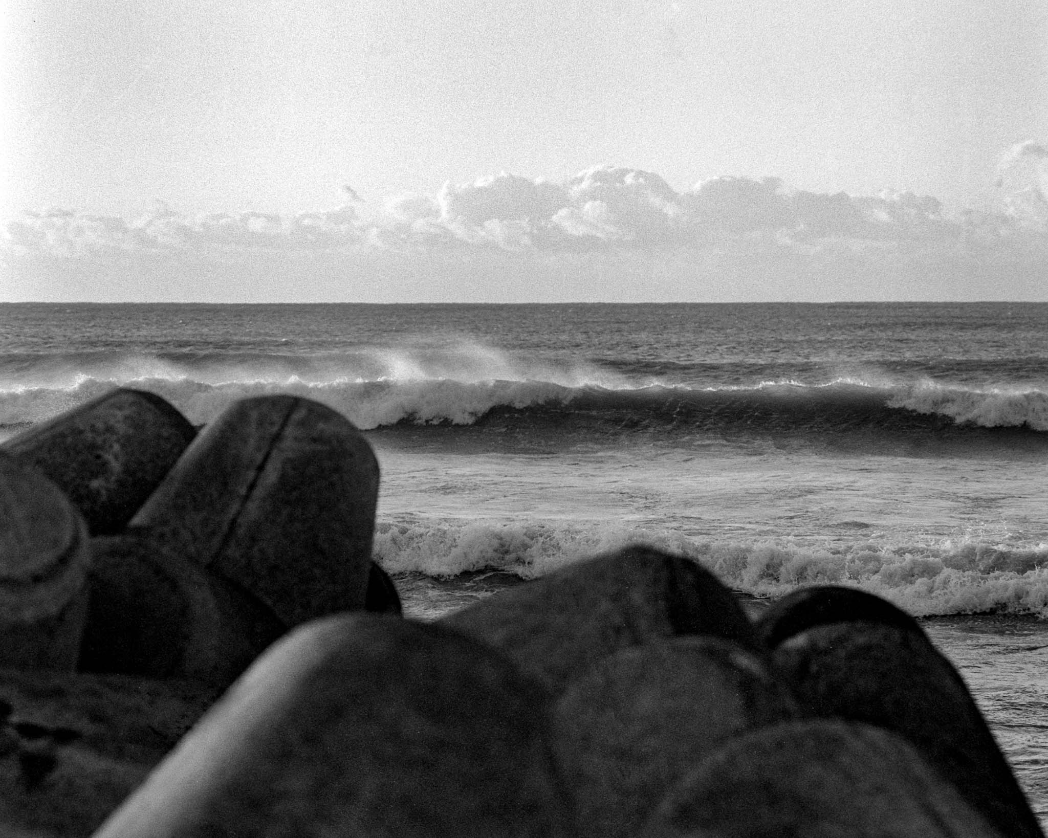Monochrome photograph of a serene coastline featuring eroded boulders, dynamic ocean waves, and dramatic clouds.