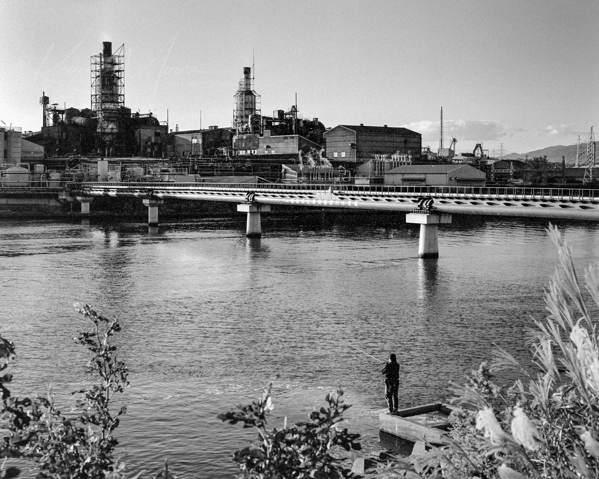 Solitary figure fishing in calm river under an industrial mid-20th century bridge, black and white photo.