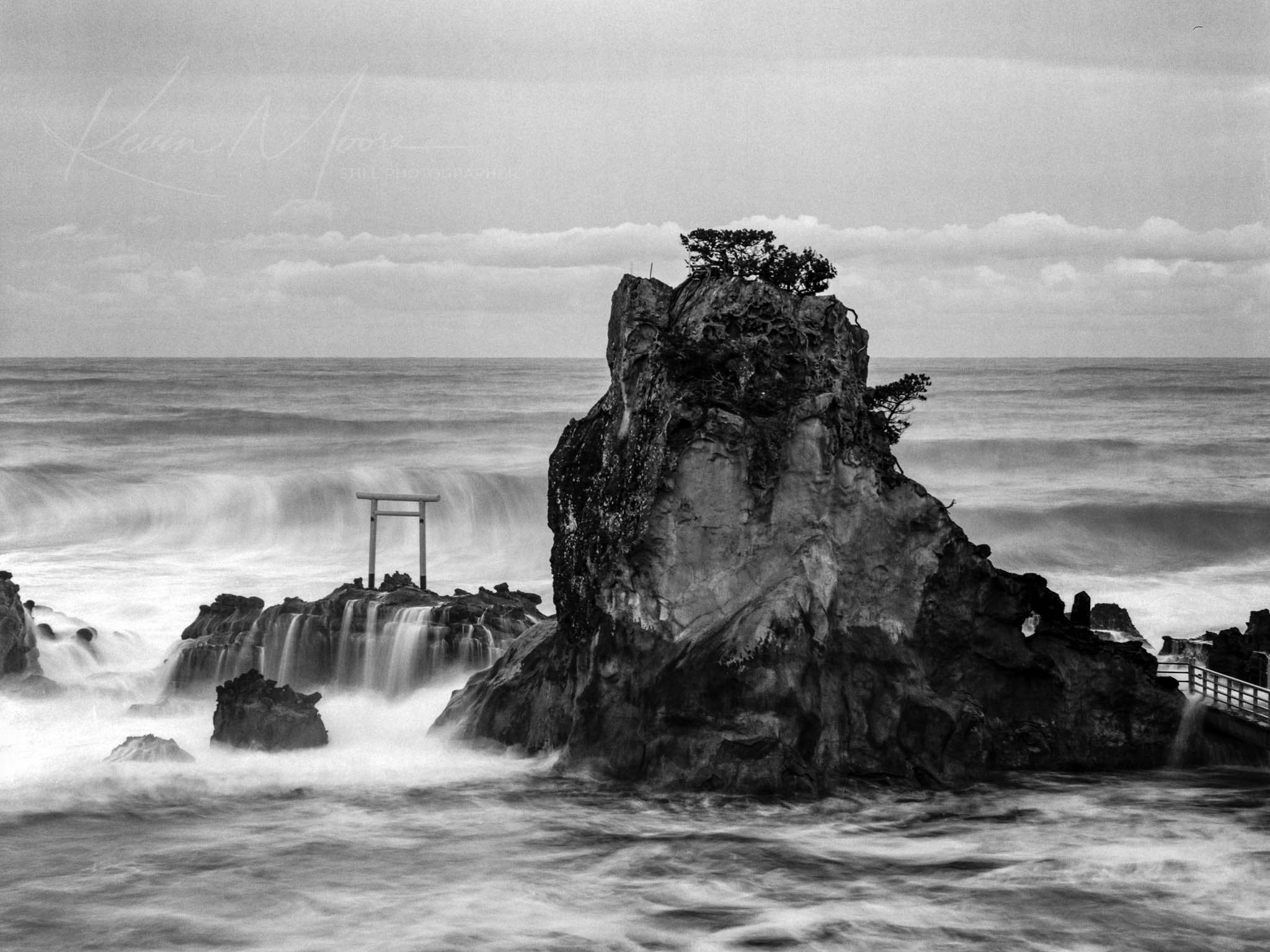 Monochrome image of rugged rock formation and Shinto shrine amidst turbulent seas.