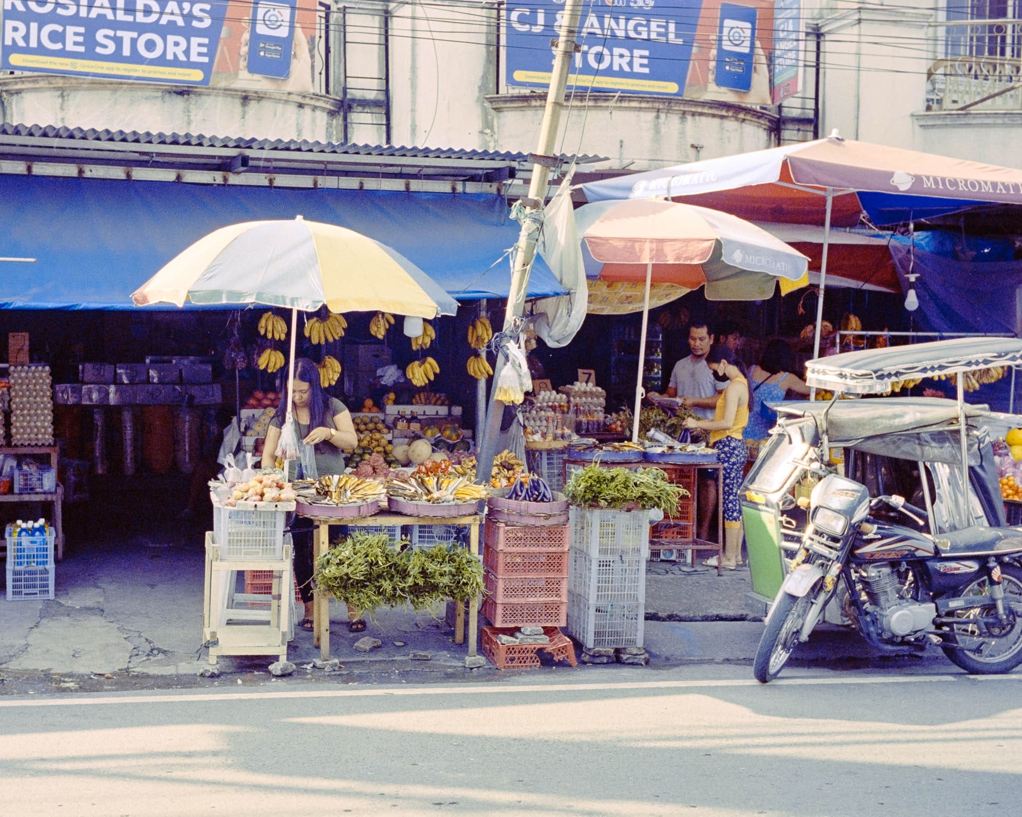 Philippines street market scene with colorful fruit stalls, parked tricycle, and local shops under clear sky.