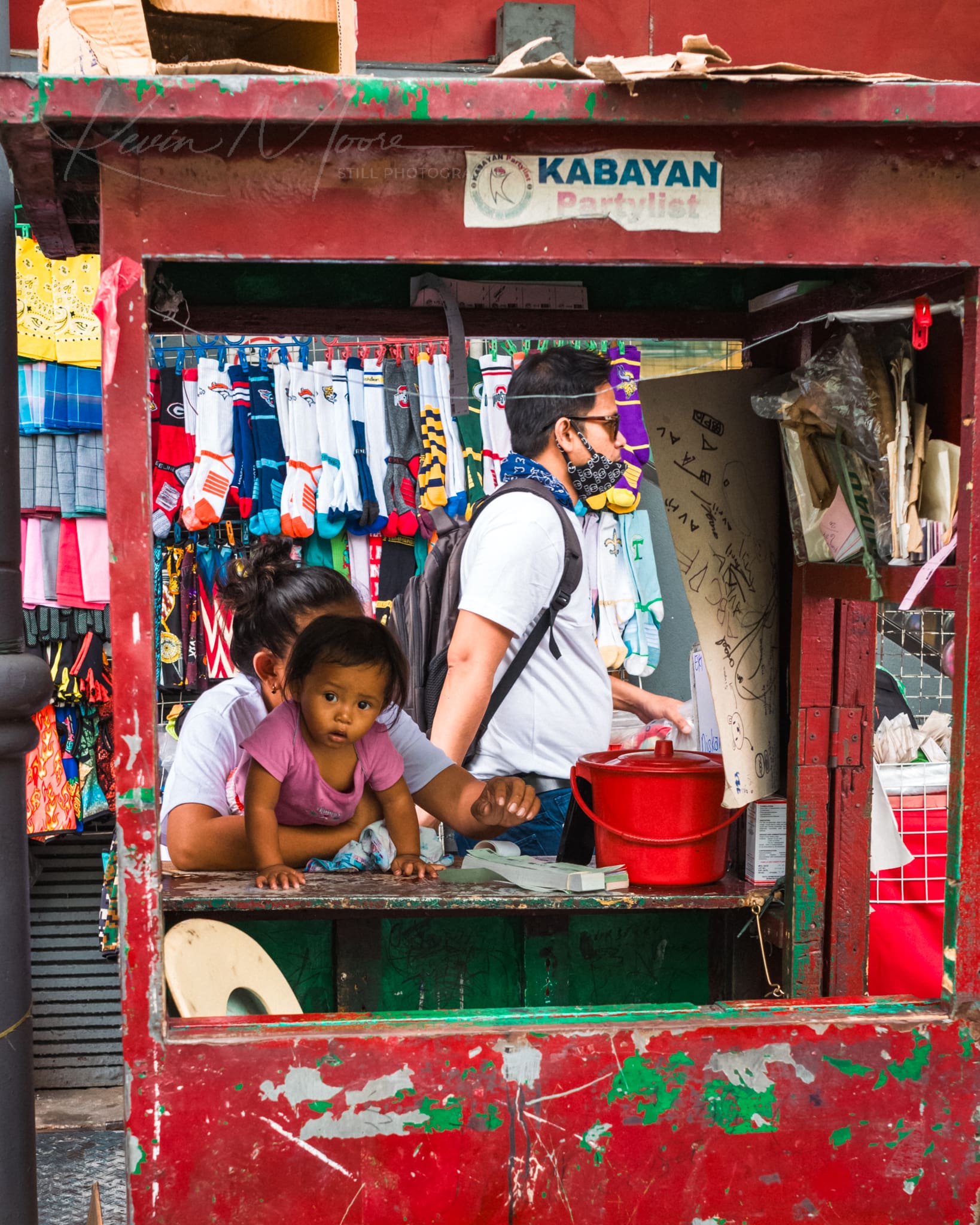 Filipino street vendor stall with a toddler and colorful merchandise during COVID-19.