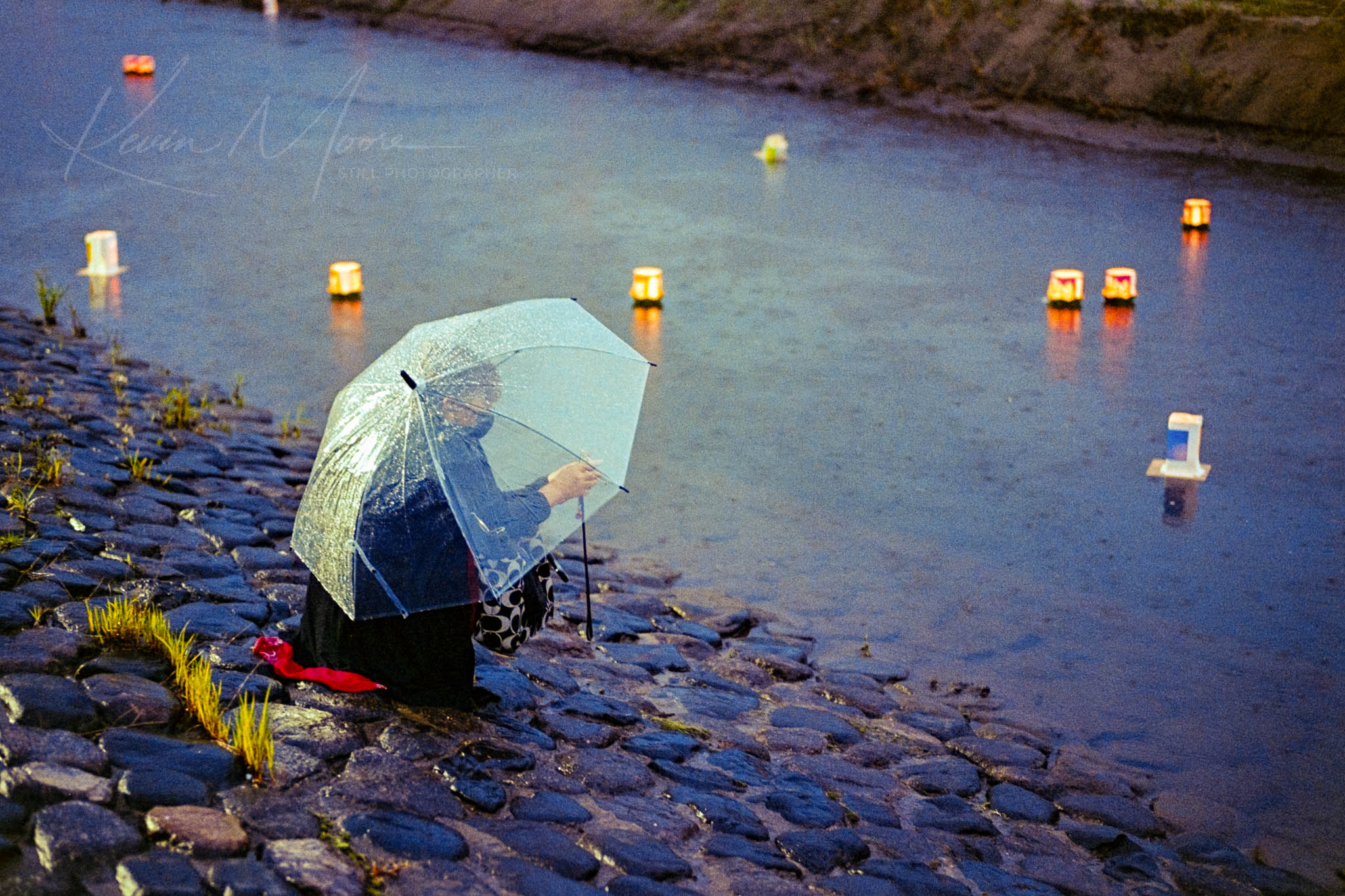 Woman under umbrella releasing glowing lanterns on cobblestones near tranquil waters at twilight.