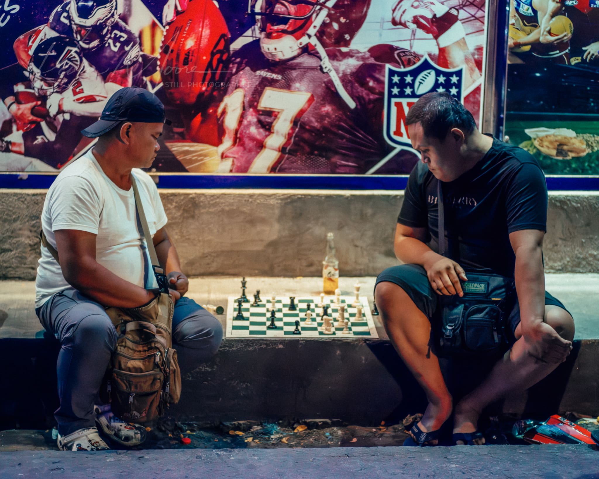 Two men engrossed in an outdoor street chess game at night with a vibrant NFL mural backdrop.