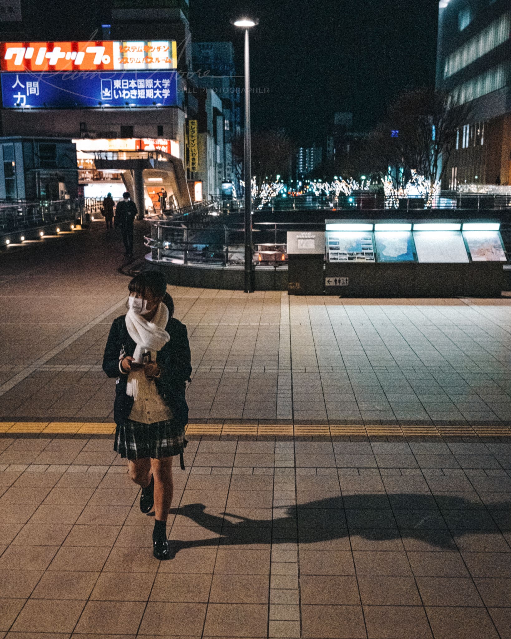 Masked Japanese school girl using smartphone in vibrant, illuminated commercial district in Japan at night.