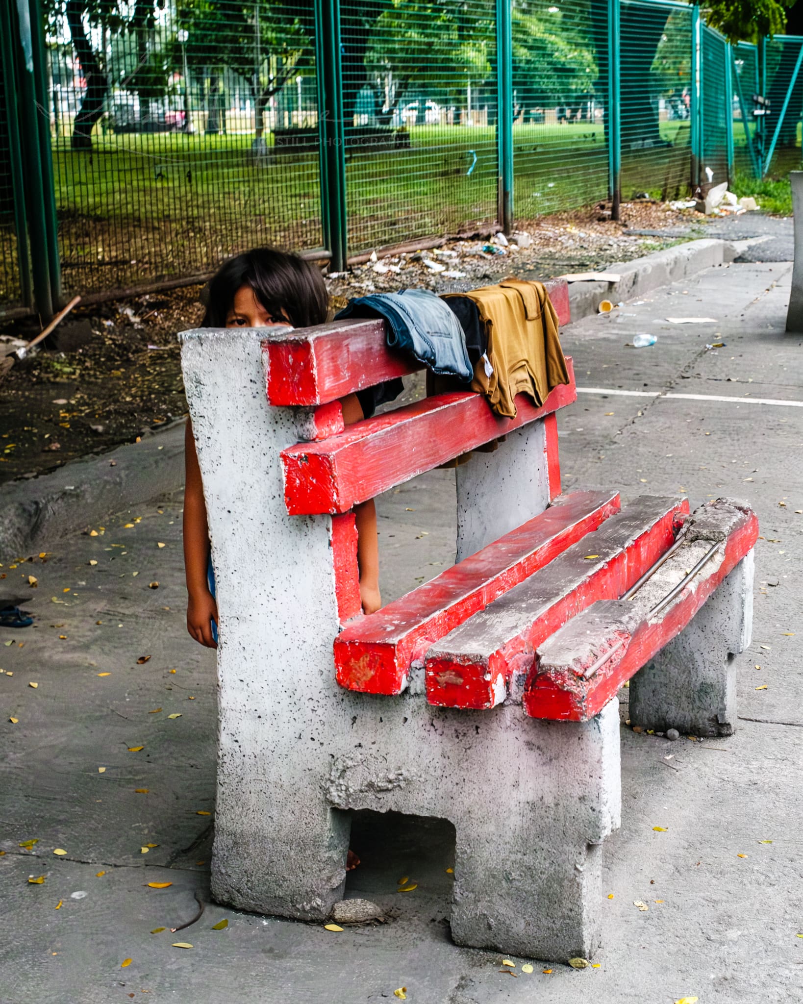 Child peeking from behind a neglected urban park bench with clothes draped over it.