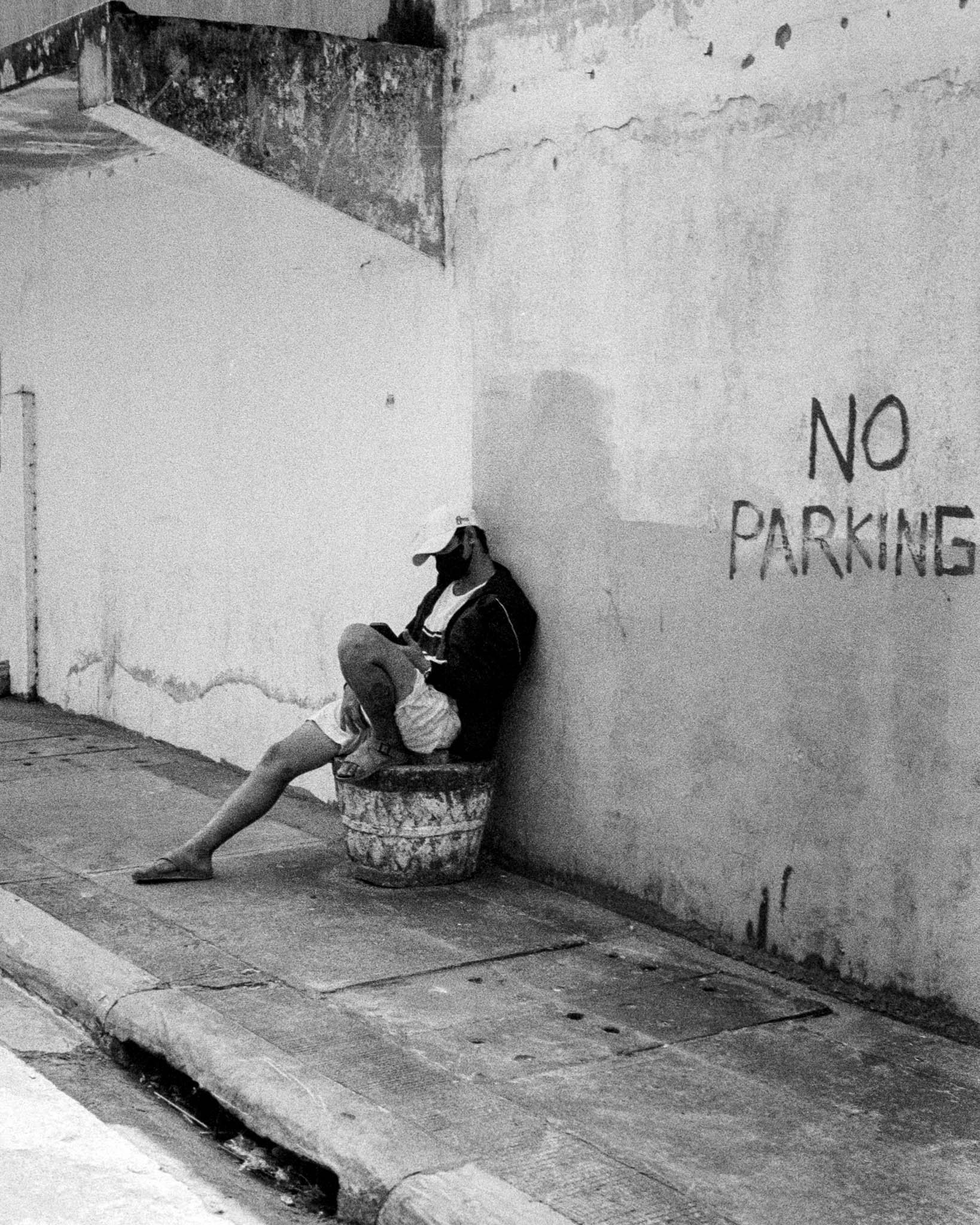 Black and white photo of lone individual seated under a NO PARKING sign, suggesting urban defiance.
