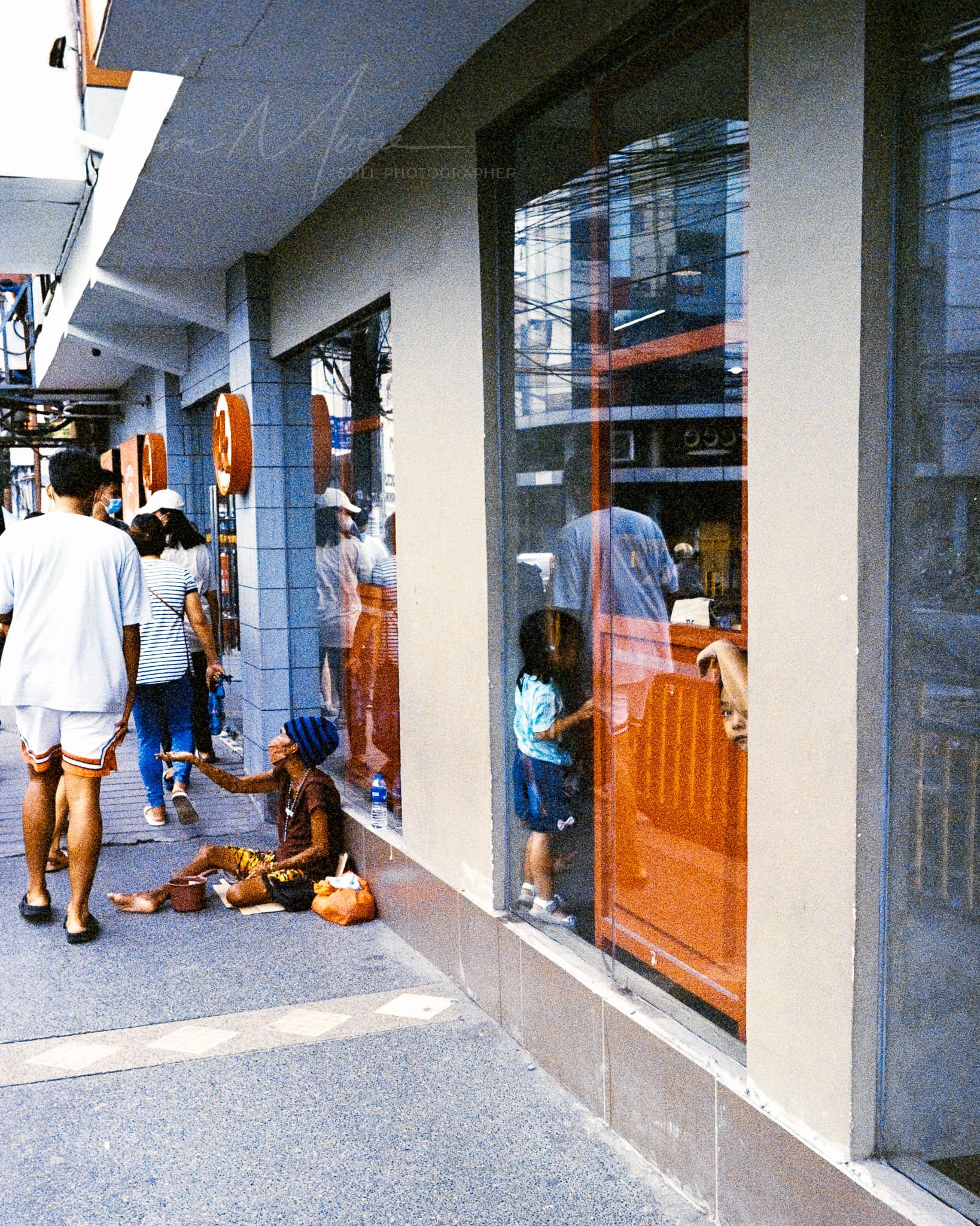 Busy urban scene depicting contrast between retail bustle and visible poverty on the sidewalk.