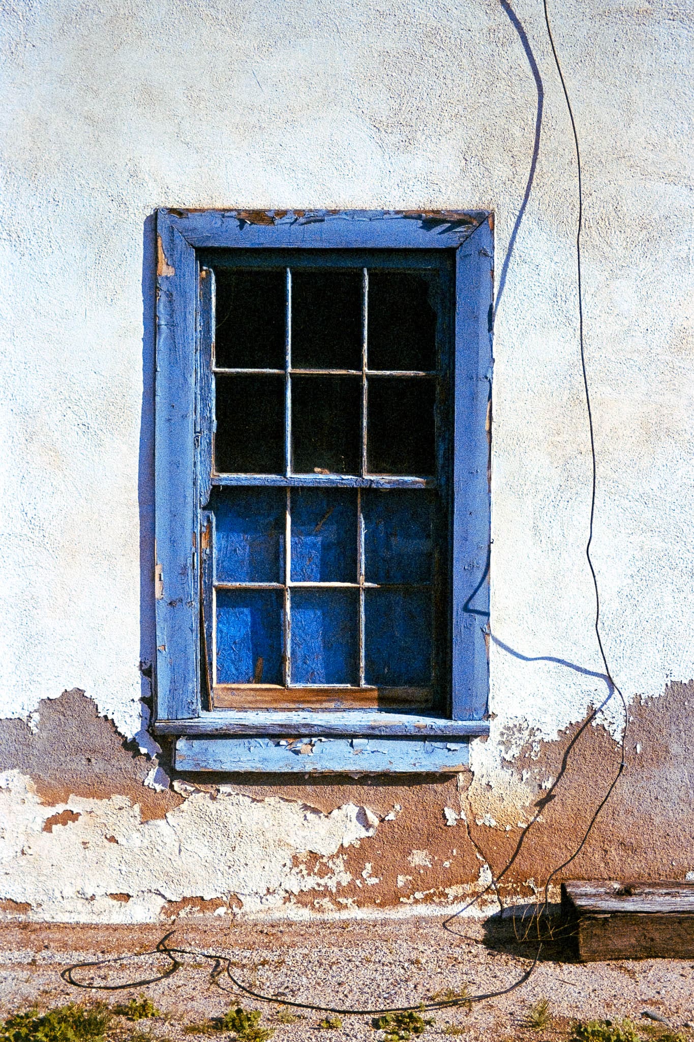 Daytime view of a rustic, faded blue window on a decaying wall with a single black wire.